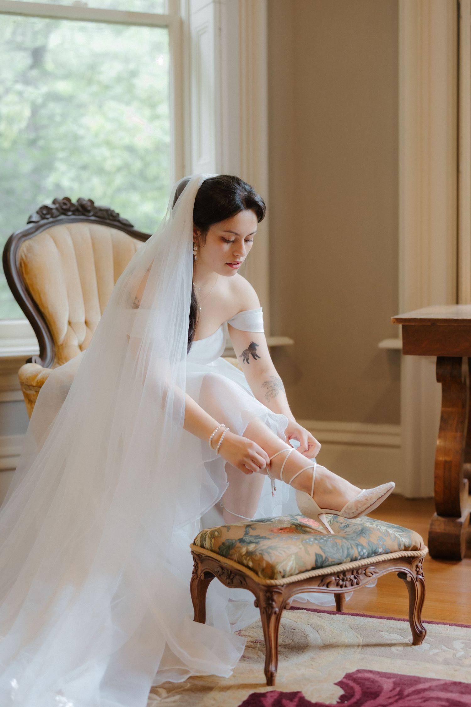 Elegant bride in white gown and veil laces up heels while seated in vintage chair by window.
