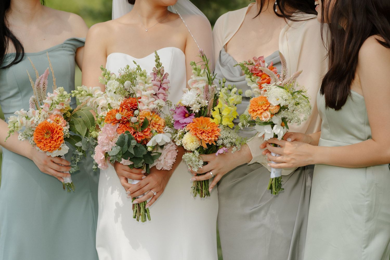 Bride and bridesmaids in sage green dresses holding colorful wildflower bouquets with orange, pink, and white blooms.