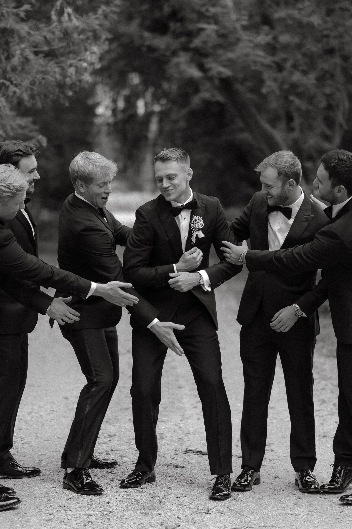 Groom surrounded by smiling groomsmen in black tuxedos sharing a joyful moment outdoors in a black and white wedding photo.
