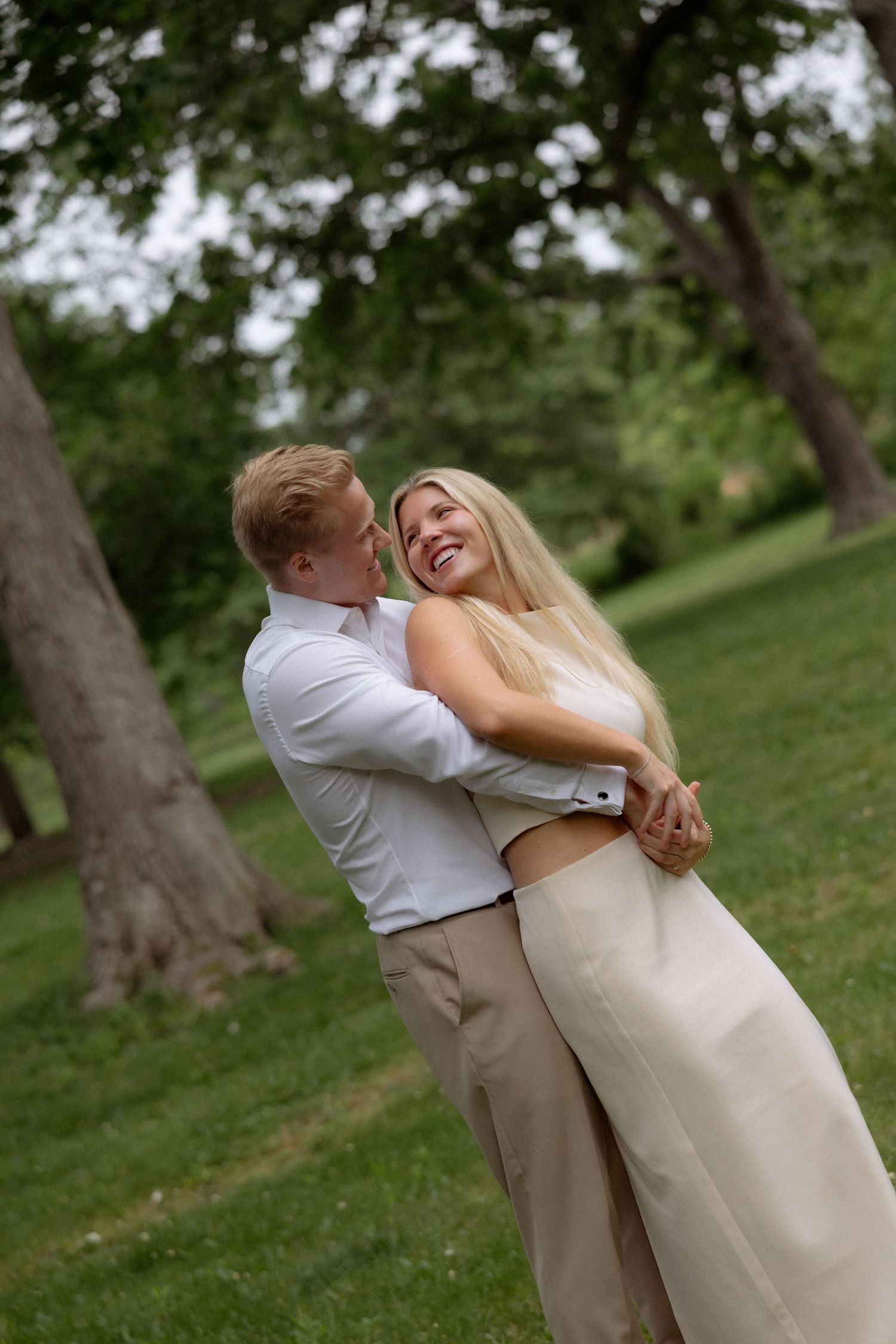 A joyful couple embraces and laughs together in a green park, man in white shirt holding woman in cream skirt.