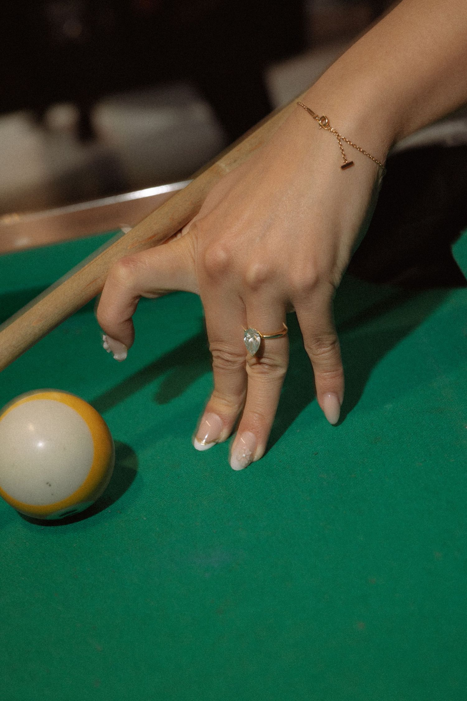 Close-up of a woman's hand with a diamond ring and gold bracelet positioning a billiard cue on a pool table.