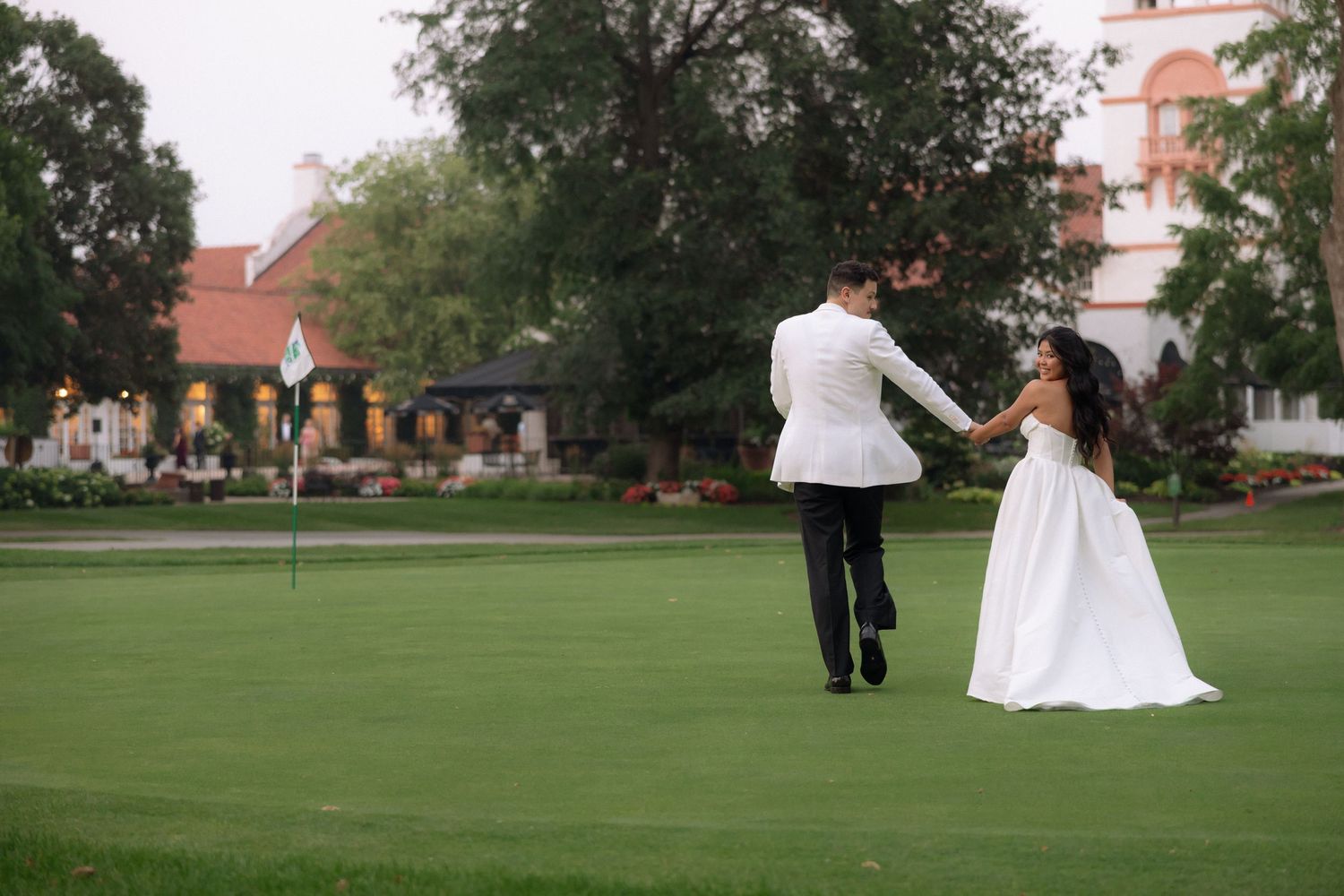 Bride and groom hold hands walking on golf course green with elegant venue in background.