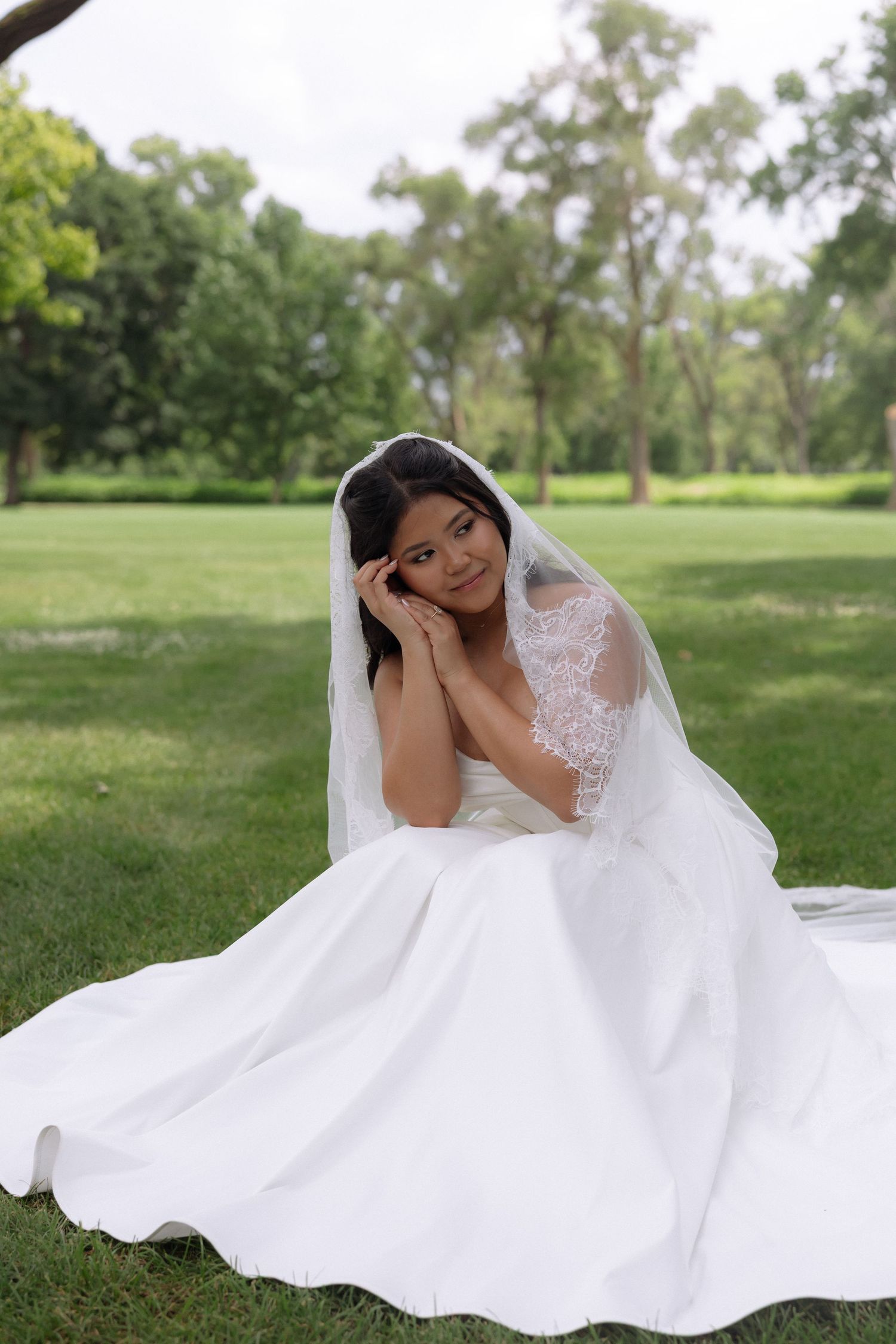 Bride in white gown and lace veil sits gracefully on green grass in a park setting.