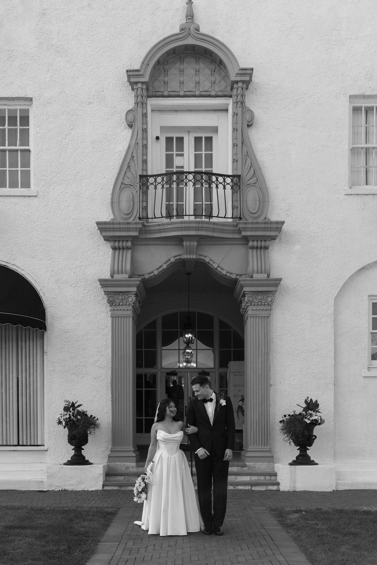 Bride and groom stand before ornate white building entrance in elegant black and white photo.