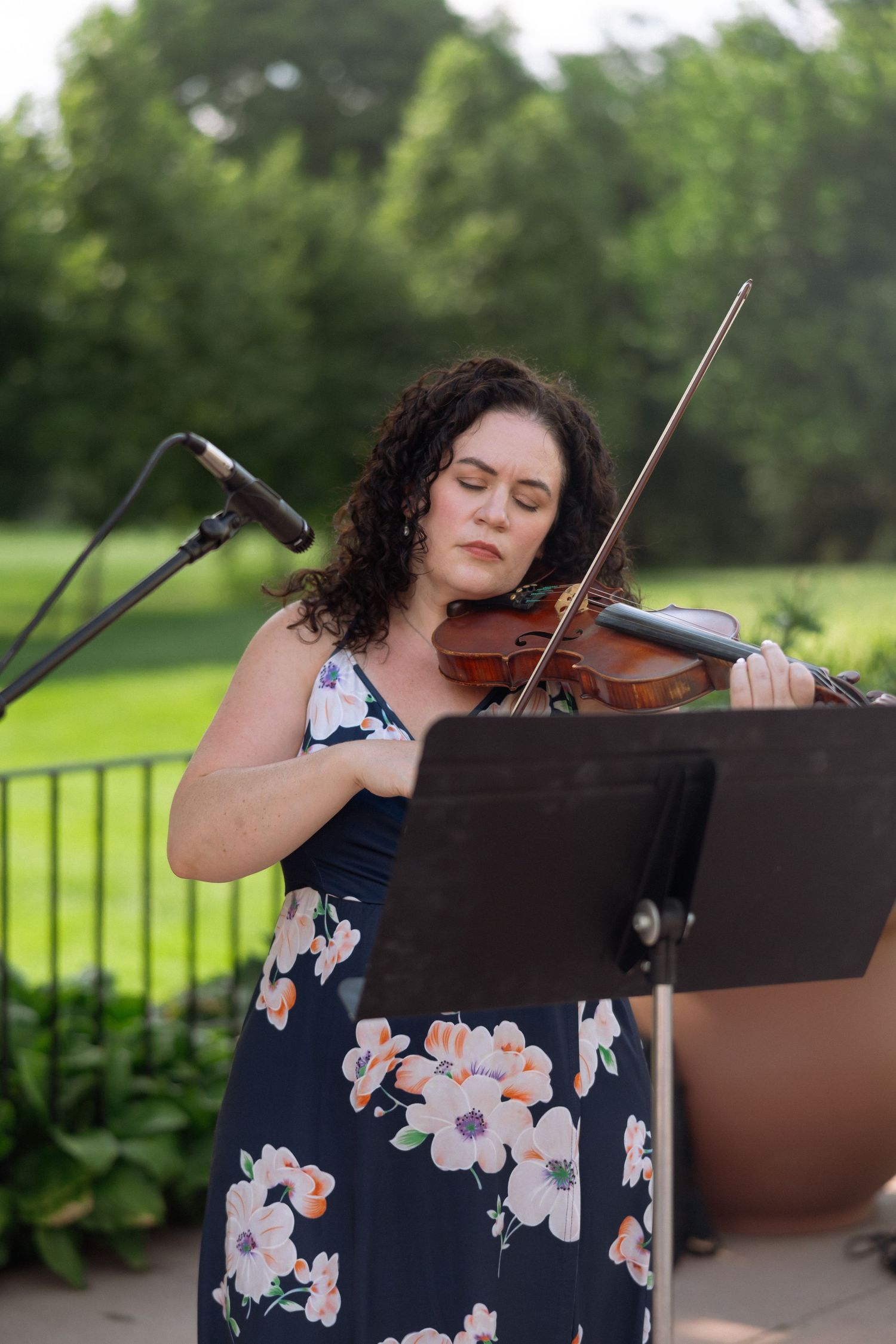 Woman in floral dress playing violin outdoors near microphone stand with green park background.