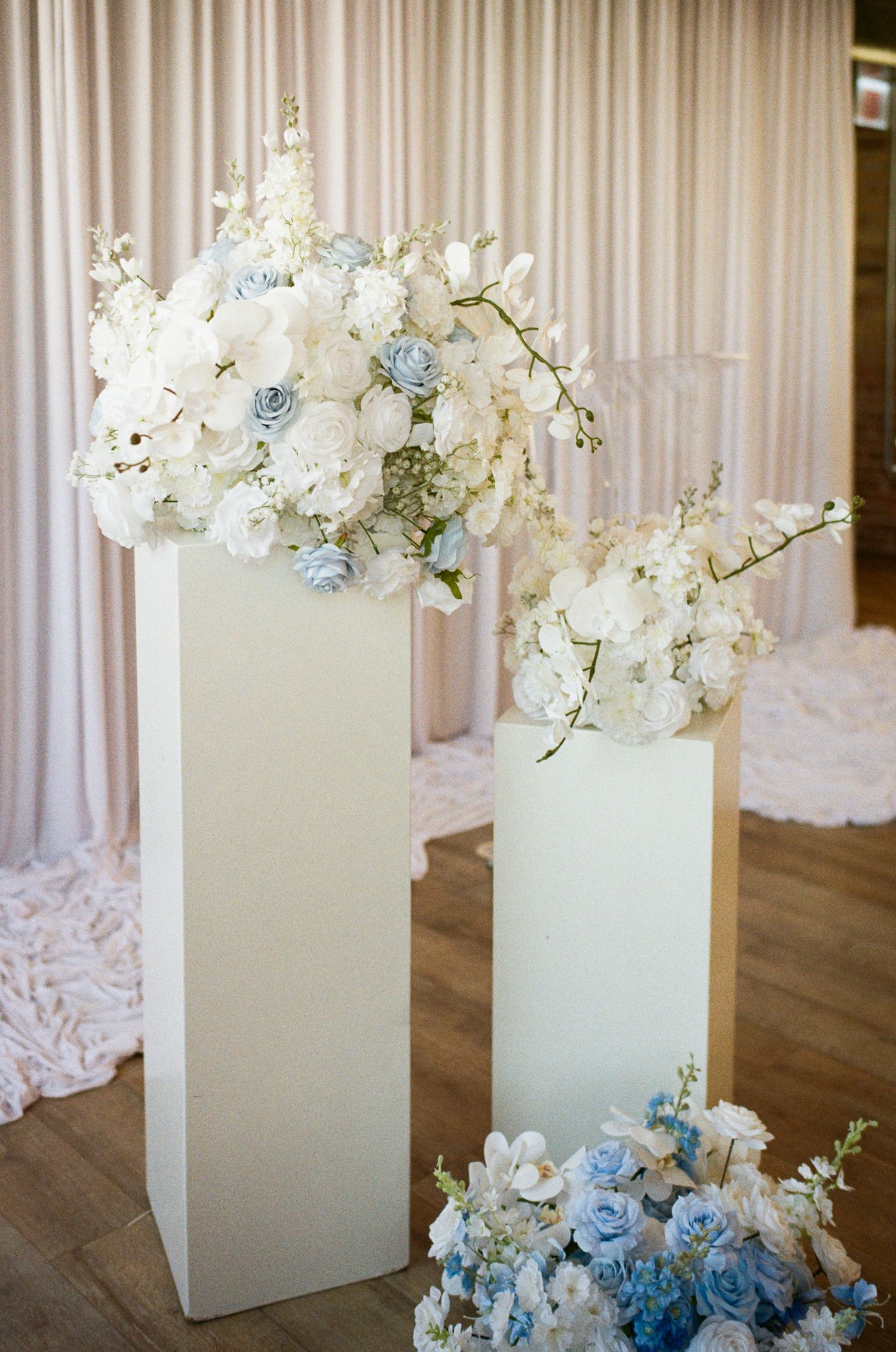 White pedestal stands topped with white and blue floral arrangements at an elegant wedding ceremony venue.