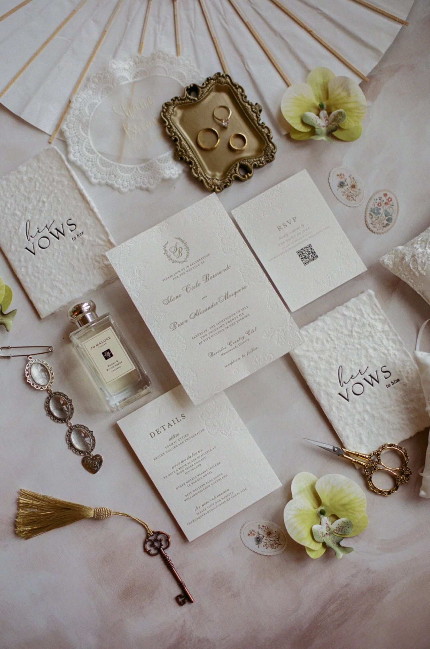 Elegant wedding flat lay with ivory invitations, vow books, gold rings, Jo Malone perfume, scissors, and green orchids.