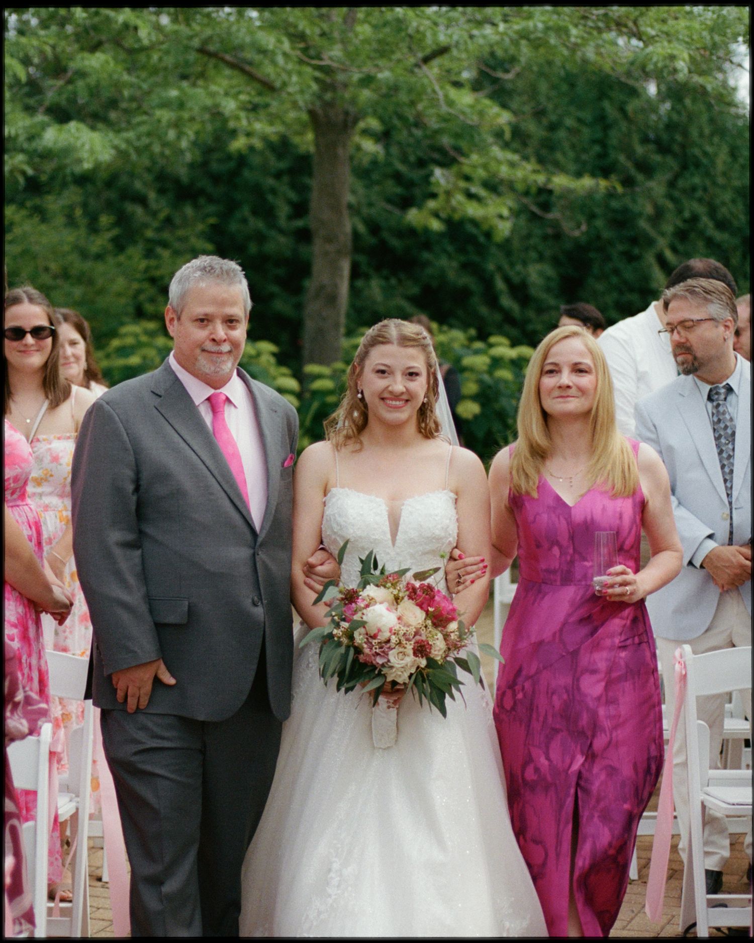 Bride in white gown walks down aisle with parents at outdoor wedding ceremony surrounded by guests.