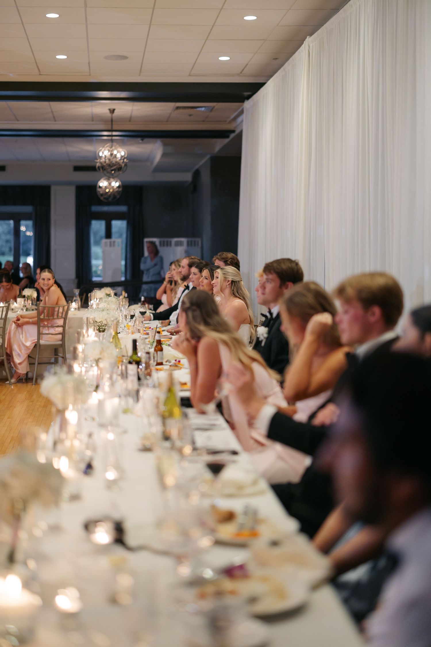 Elegant wedding reception with guests seated at a long candlelit table in a draped white venue.