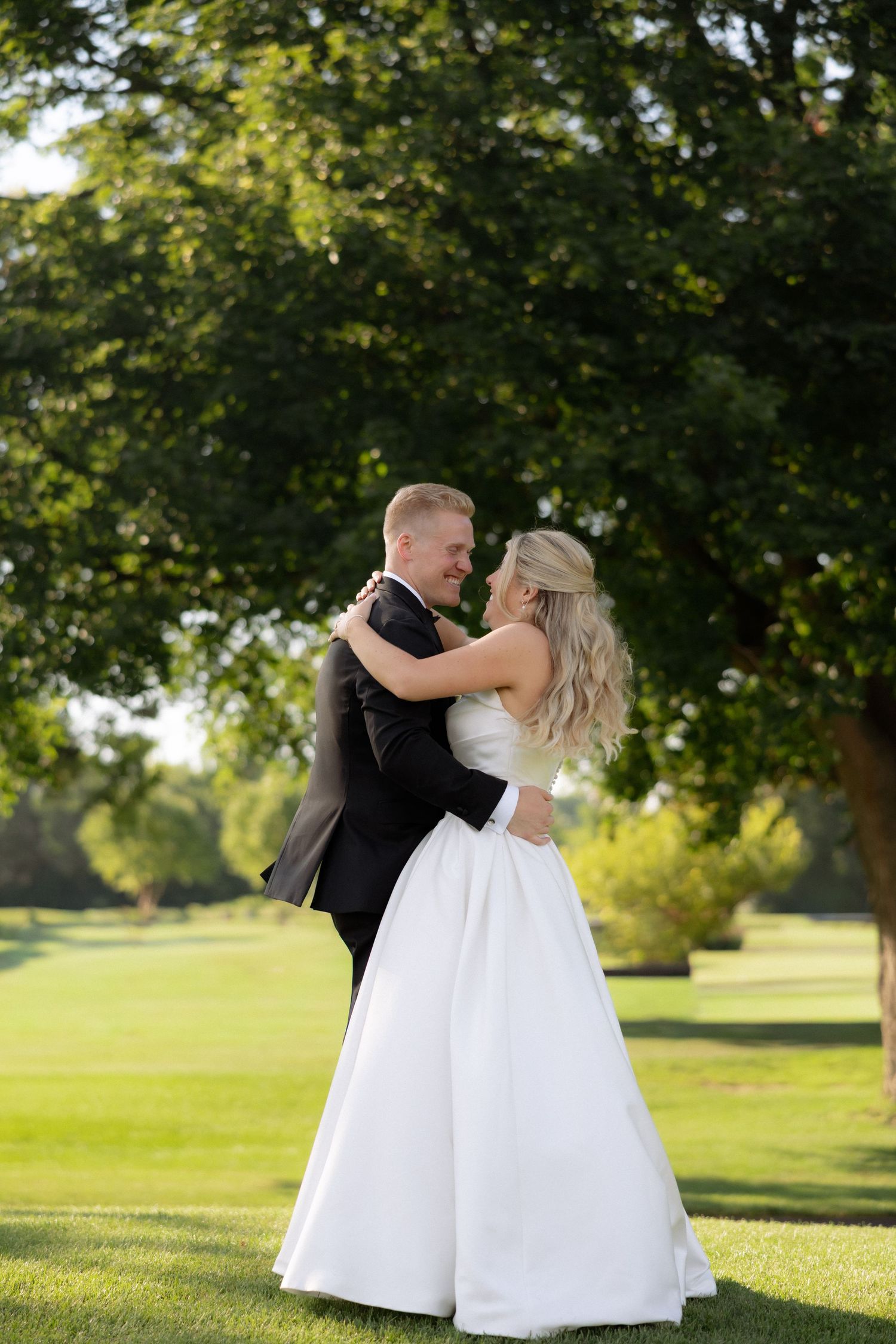 Bride and groom embrace and smile at each other on lush green lawn surrounded by trees.