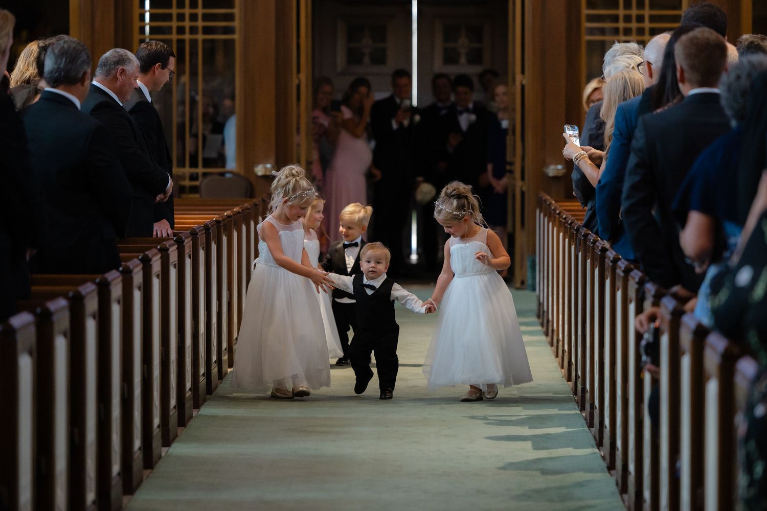 Adorable flower girls in white dresses and ring bearer in tuxedo walk down church aisle at wedding.