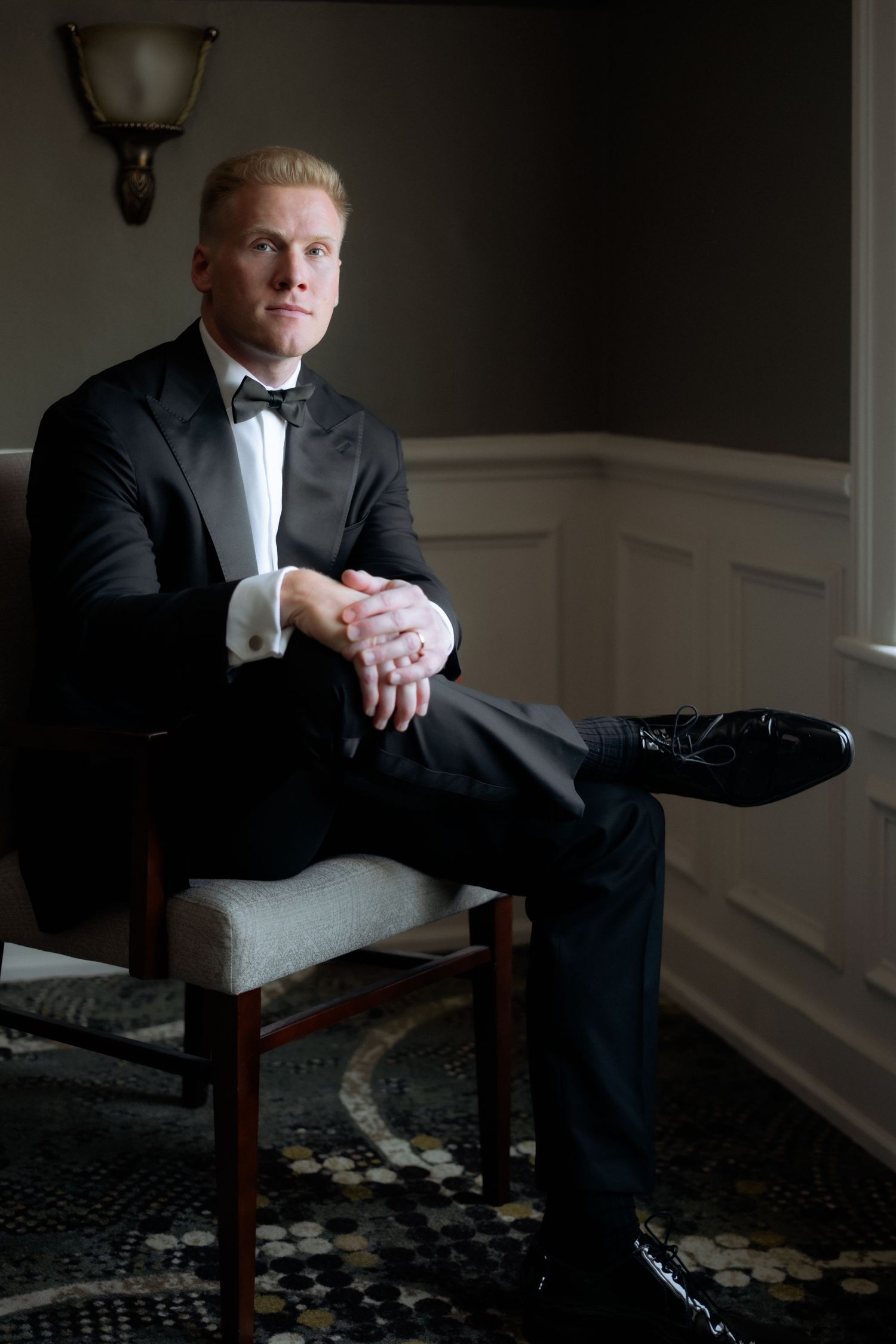 Groom in black tuxedo with bow tie sits elegantly in chair, hands clasped, in dimly lit formal room.