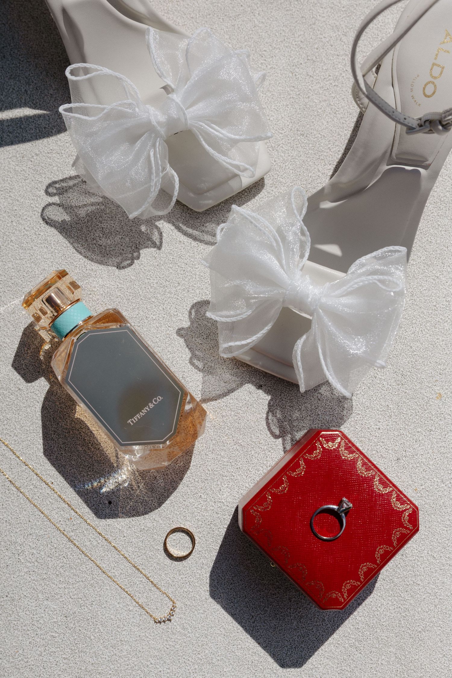 Bridal flat lay with Tiffany perfume, Cartier ring box, gold jewelry, and white bow heels on light surface.