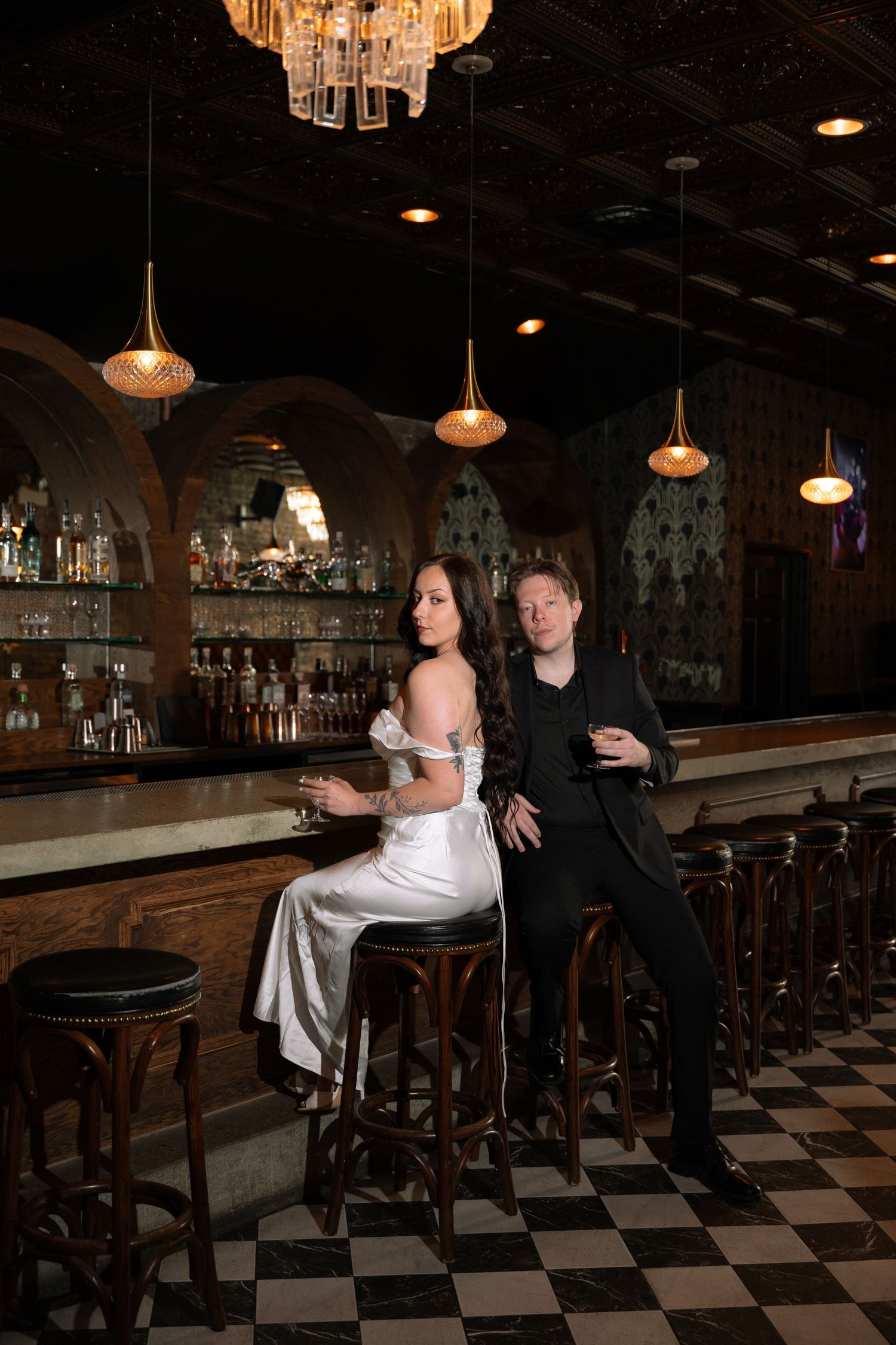 Stylish bride and groom sit at a dark, upscale bar with pendant lights and checkered floors, holding cocktails.