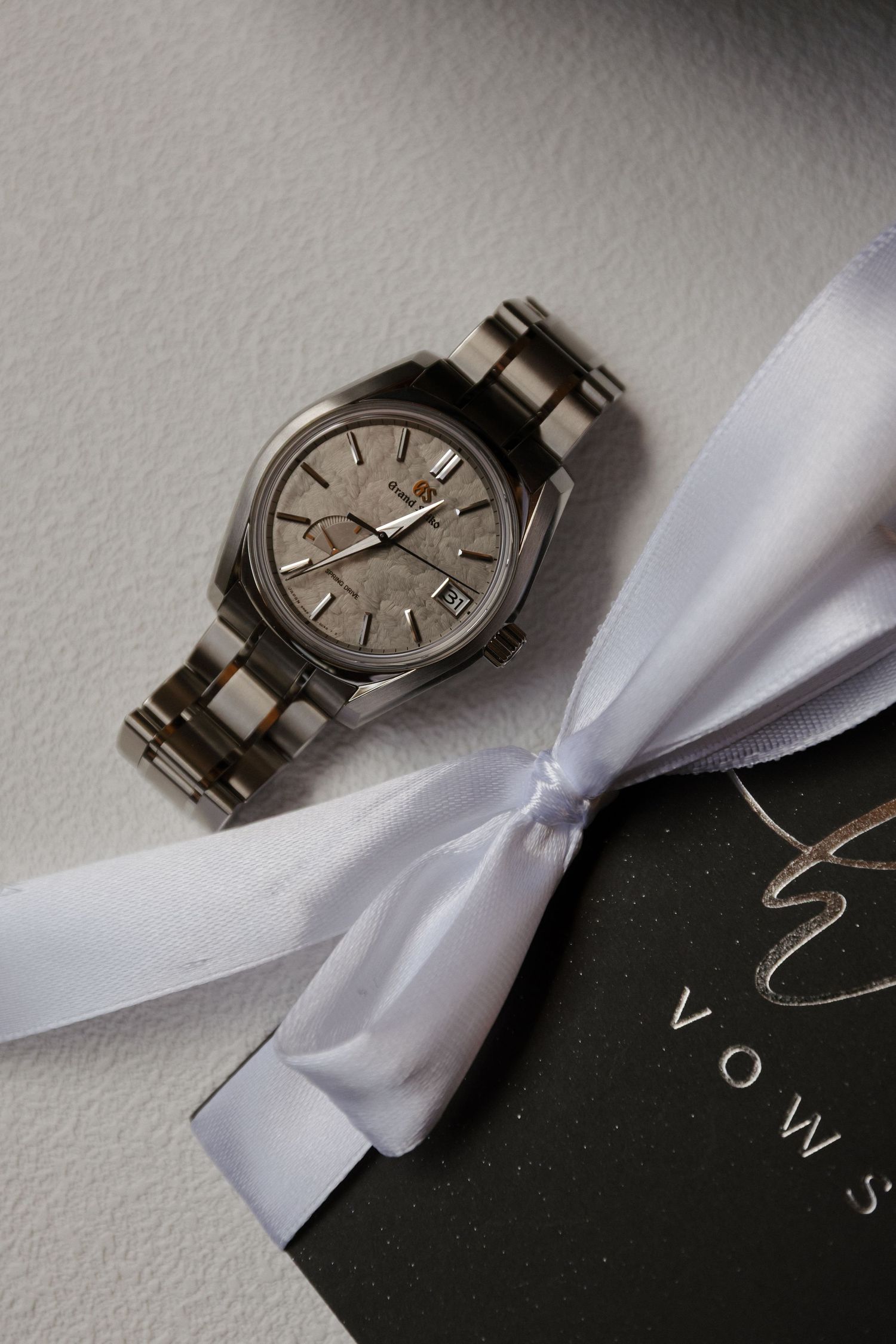 Silver Grand Seiko watch displayed on white ribbon bow atop a dark vows booklet wedding gift.