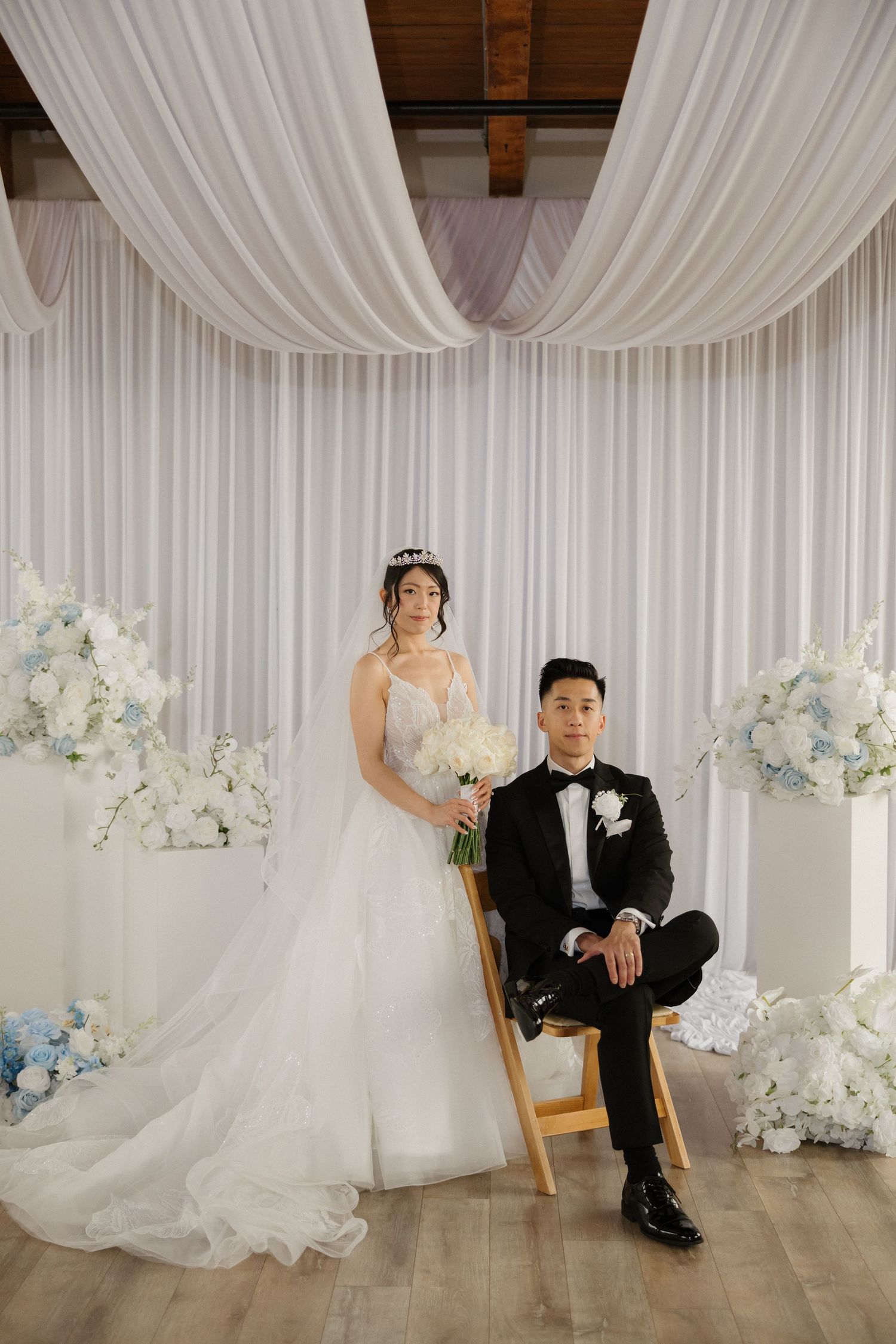 Bride in white ball gown and groom in black tuxedo pose before elegant white floral backdrop with blue hydrangeas.