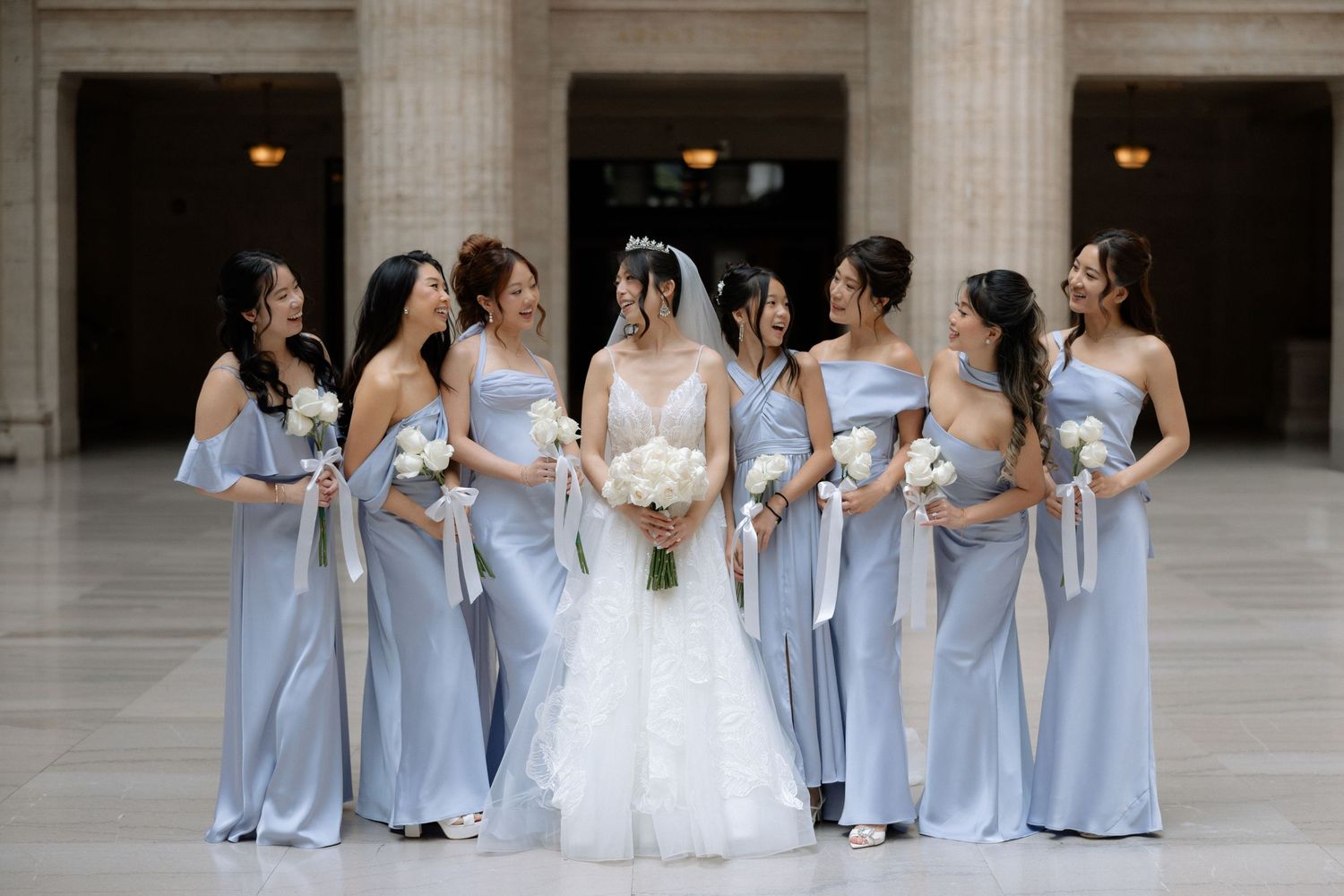 Bride in white gown poses with bridesmaids wearing matching light blue satin dresses holding bouquets.