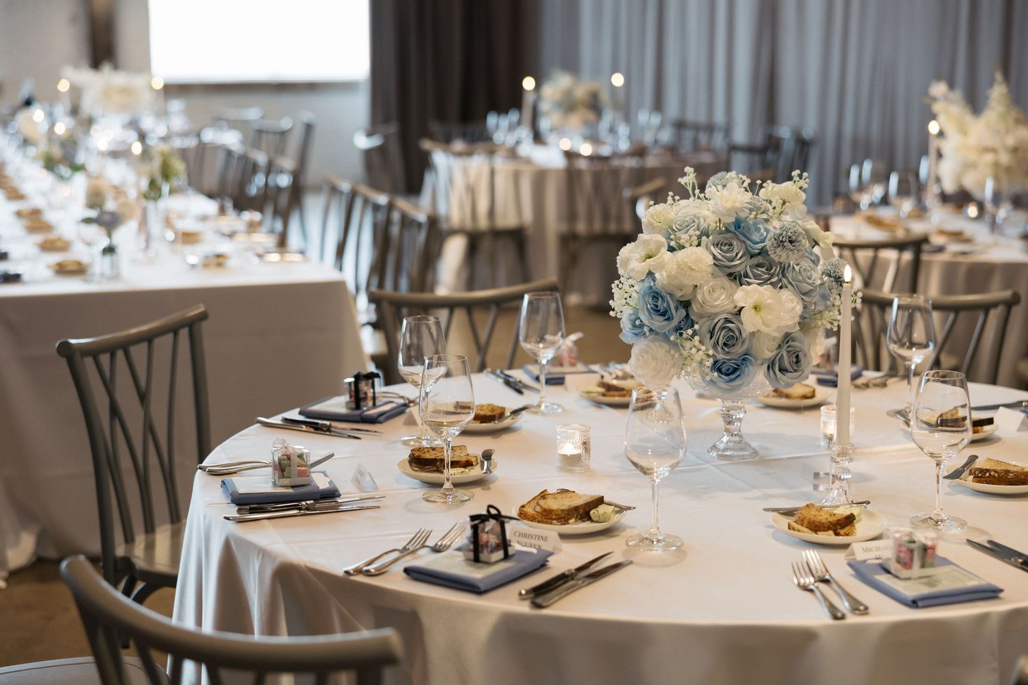 Elegant wedding reception tables set with white linens, blue and white floral centerpieces, glassware, and gold-trimmed place settings.