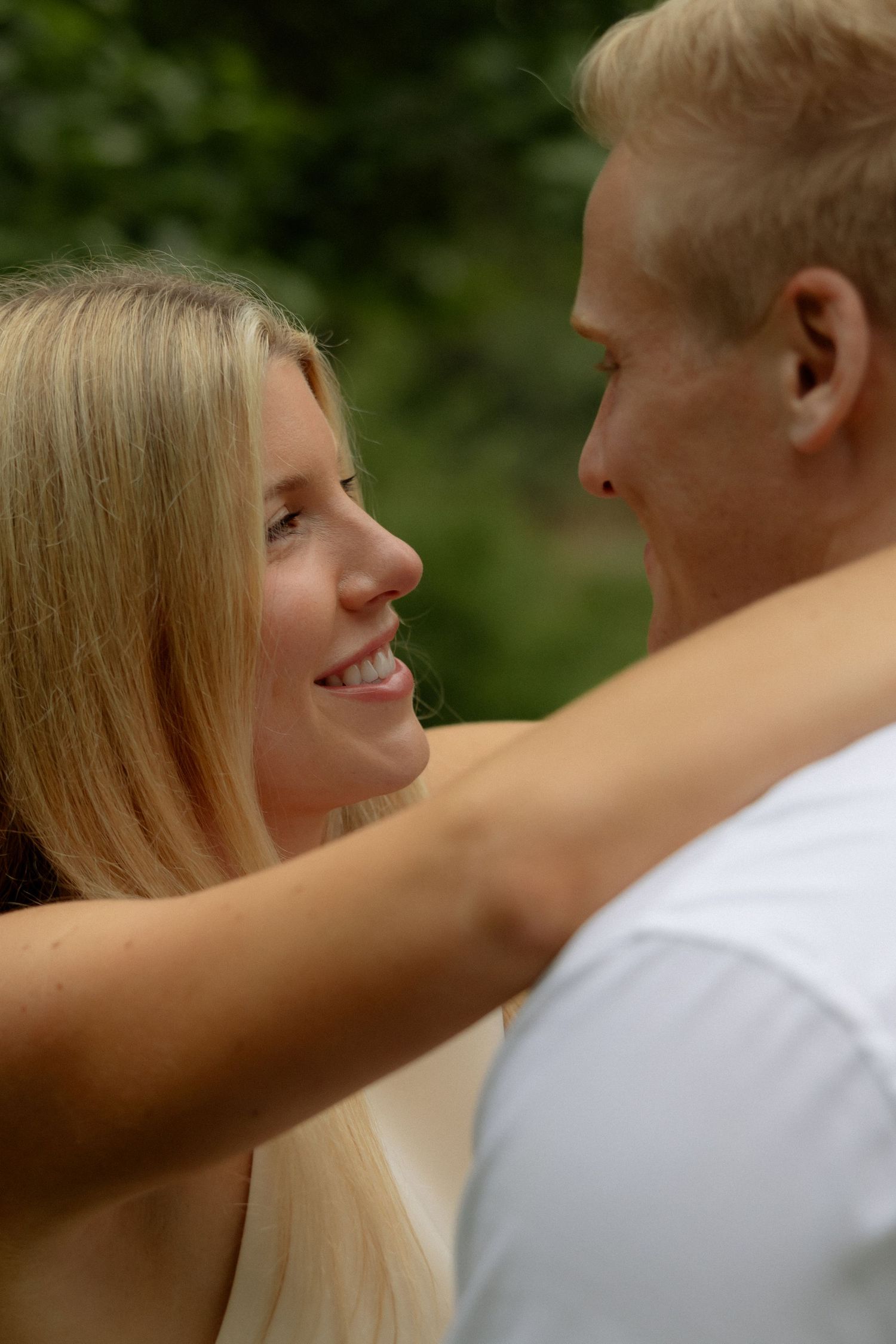 A smiling blonde woman gazes lovingly at a man while embracing outdoors with green foliage in the background.