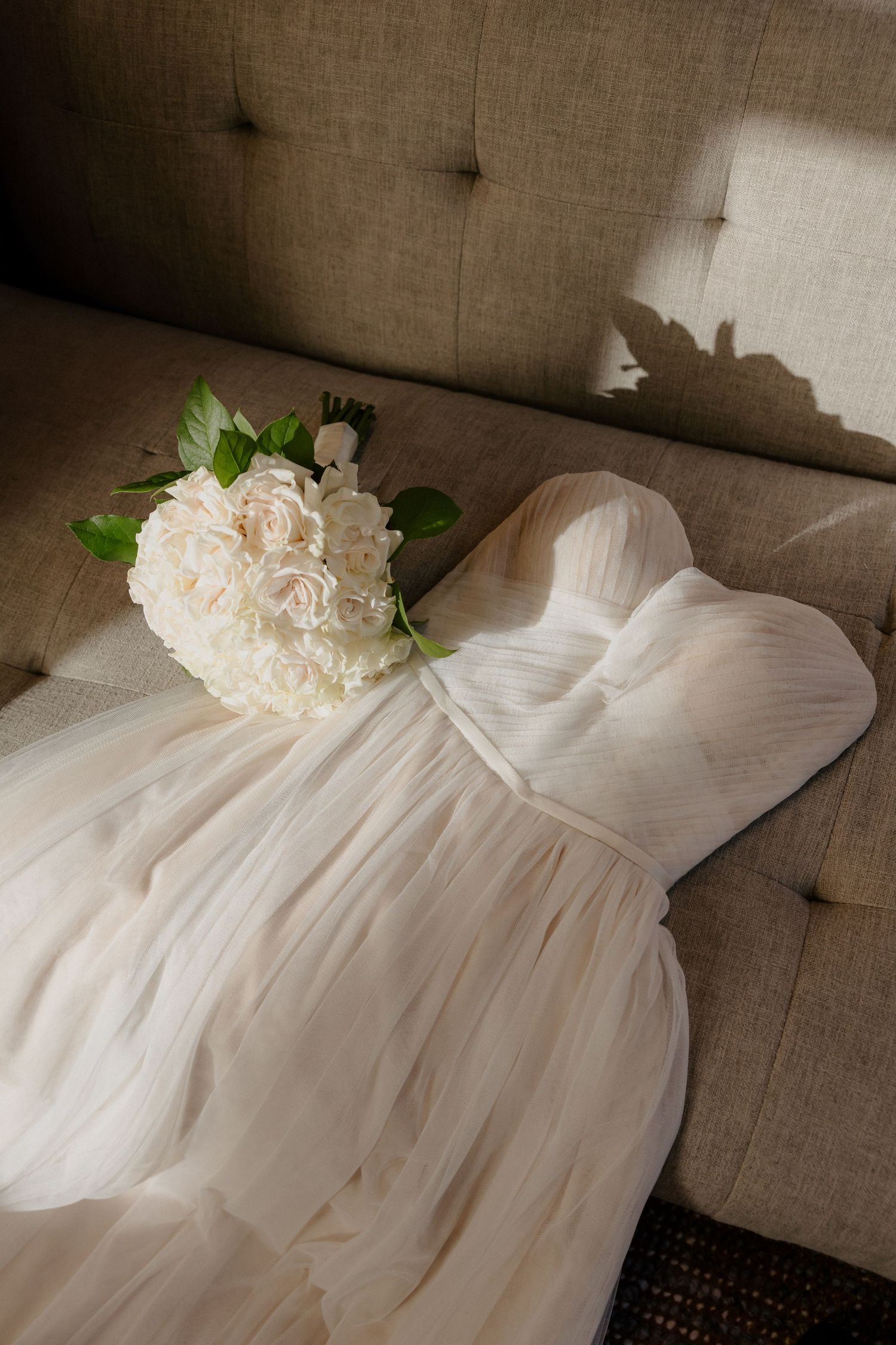 Elegant strapless wedding dress laid flat on a beige tufted sofa with a white peony bridal bouquet.