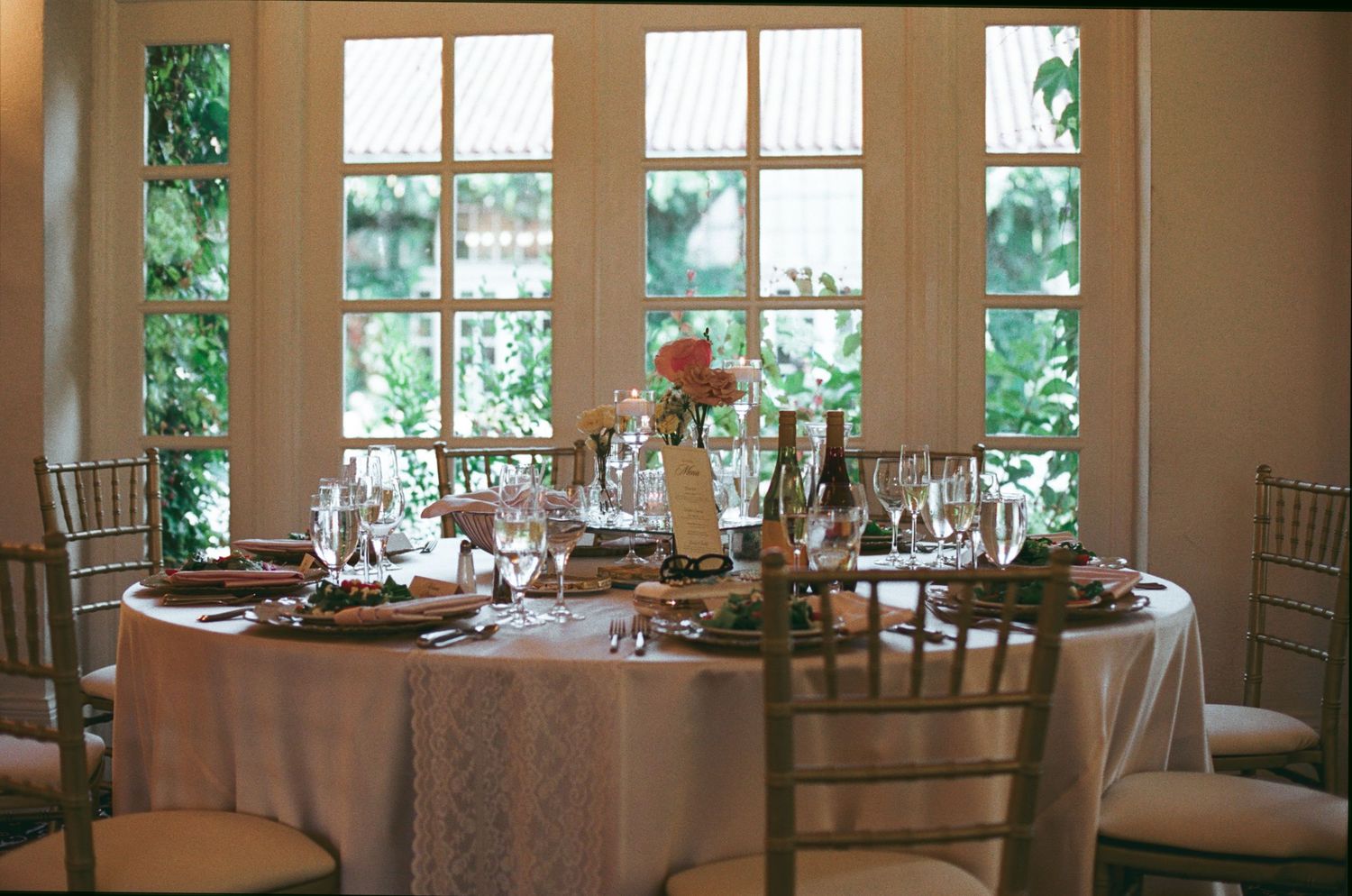 Elegant round dining table set for a wedding reception with gold chiavari chairs, wine glasses, and floral centerpiece by bright windows.