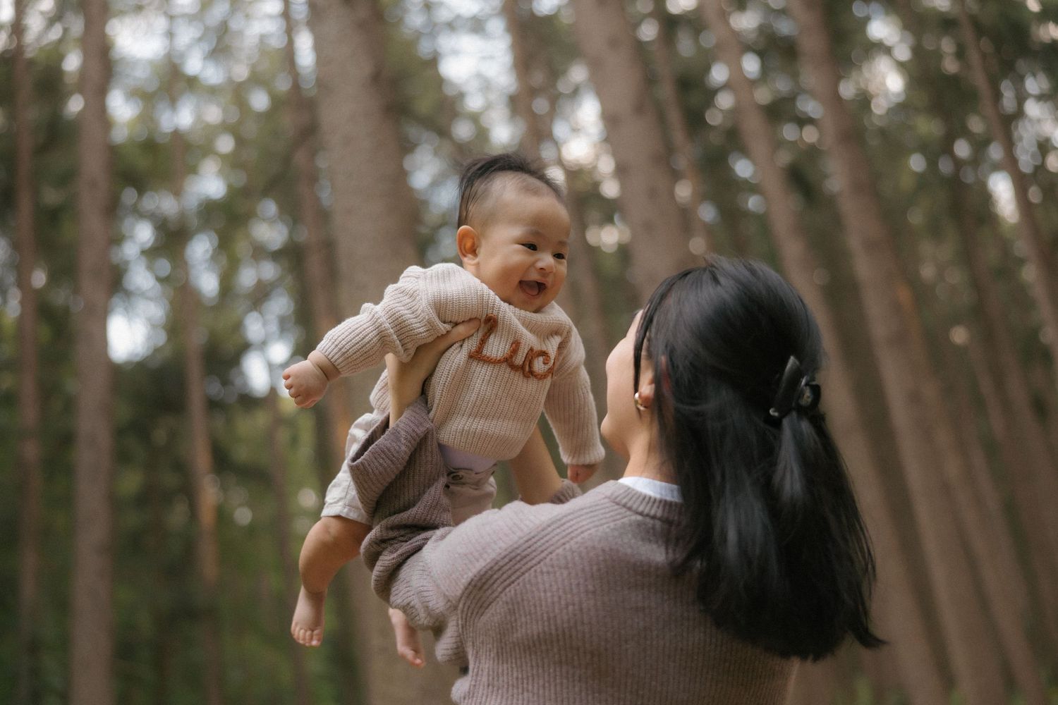 A mother joyfully lifts her laughing baby in a sunlit forest surrounded by tall pine trees.