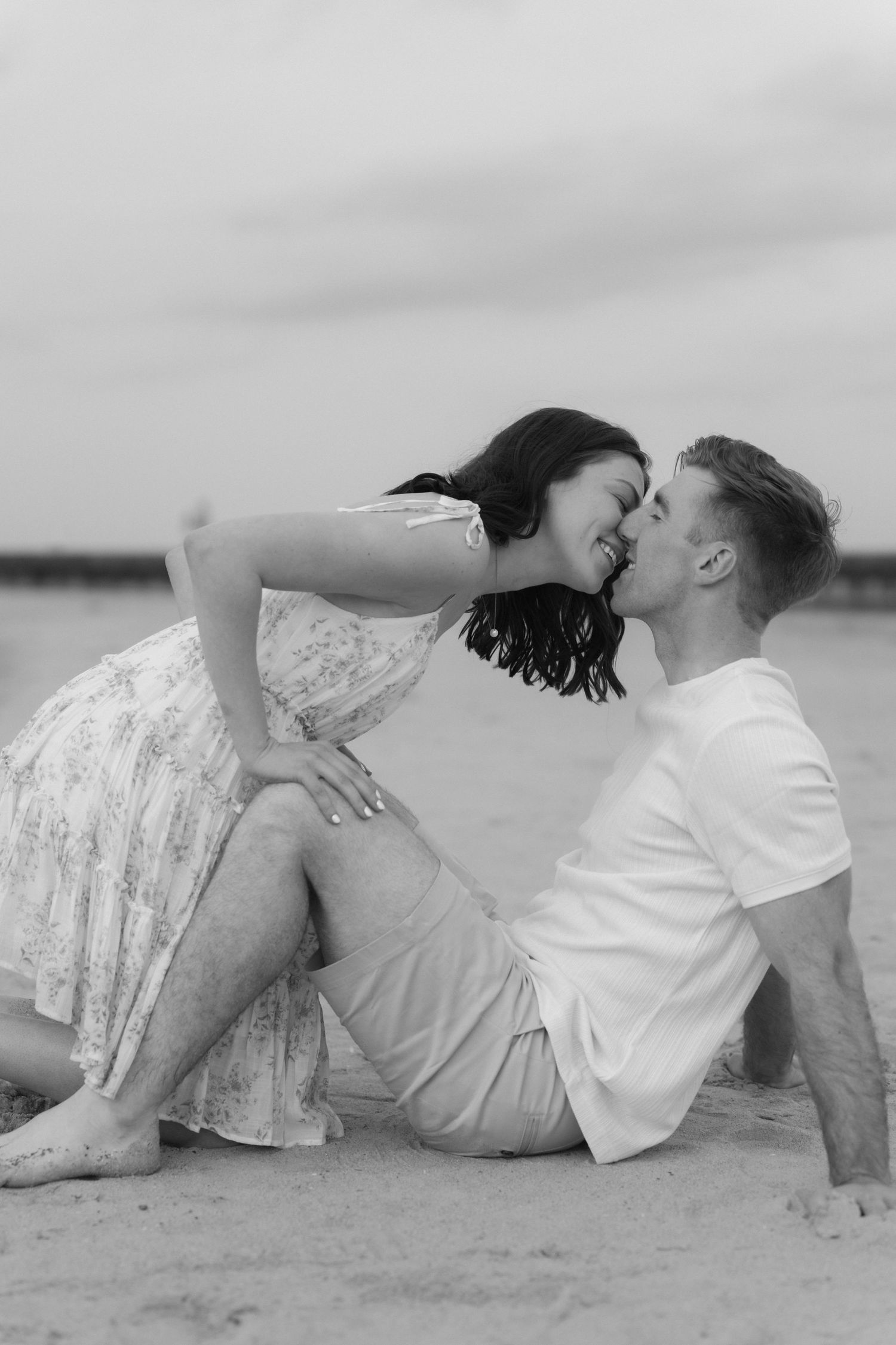Black and white photo of a couple sharing a kiss while sitting on a sandy beach, captured in a romantic outdoor setting.