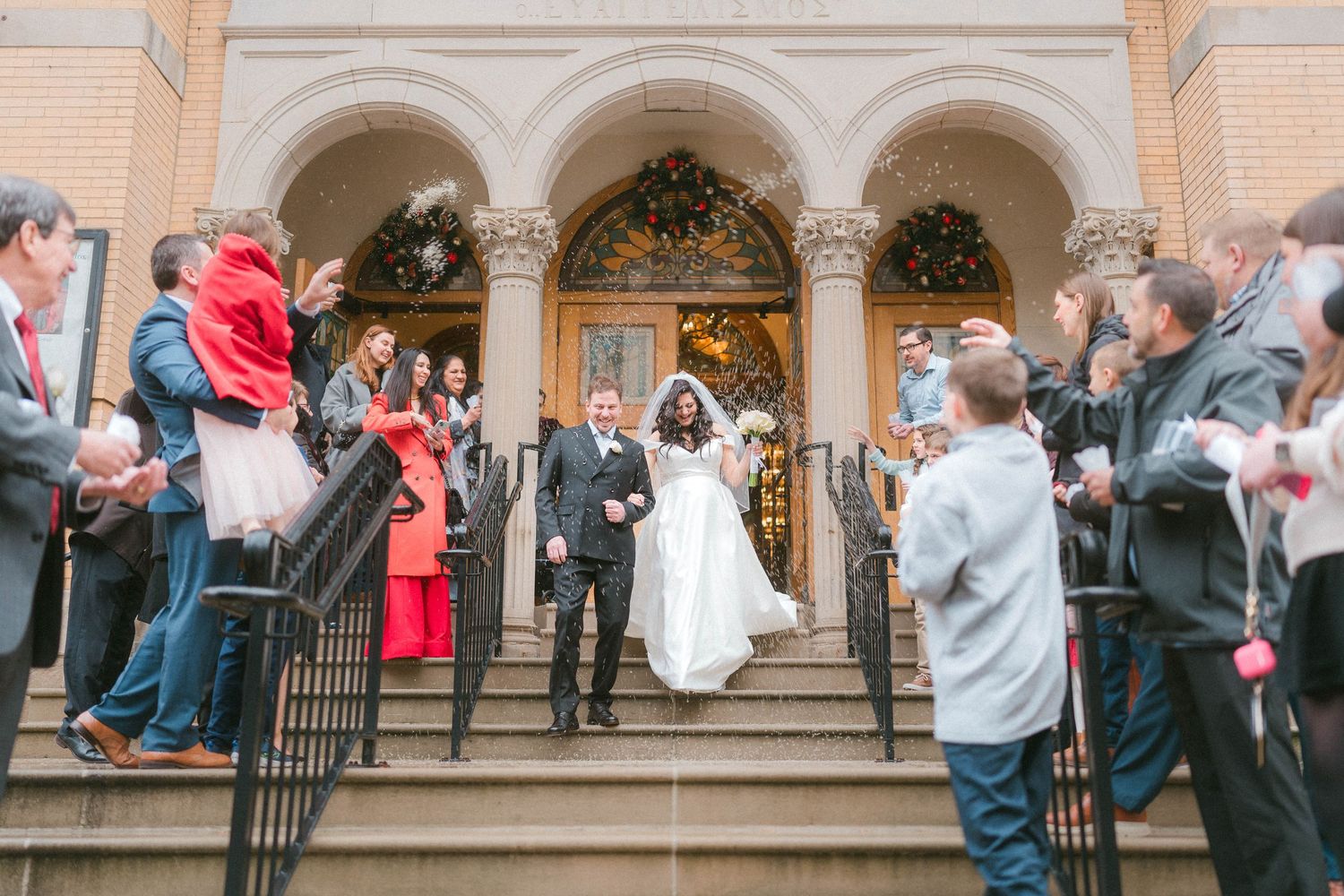 Newlyweds walk down grand church steps as wedding guests line staircase in celebration.