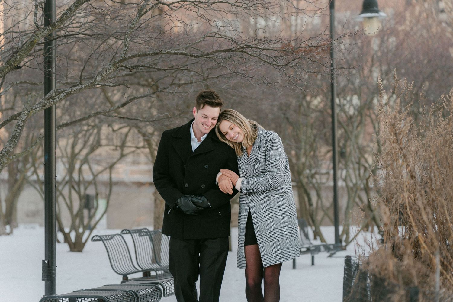 Couple walking together in snowy park, woman leaning on mans shoulder, both wearing winter coats.