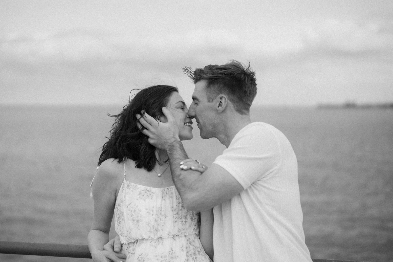A romantic black and white photo of a couple kissing near the water on a windy day.