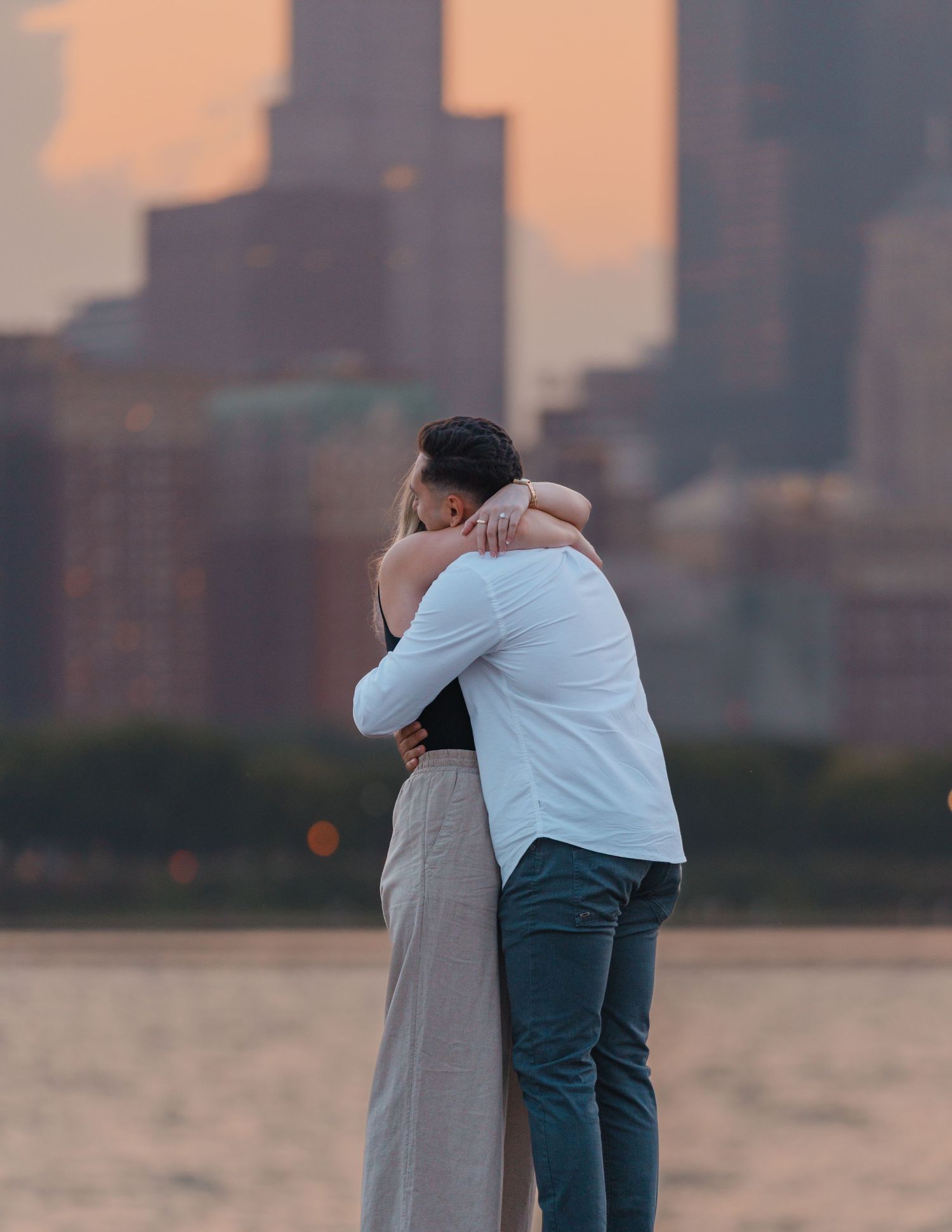 Couple embracing in a romantic hug by the waterfront with a city skyline at dusk.