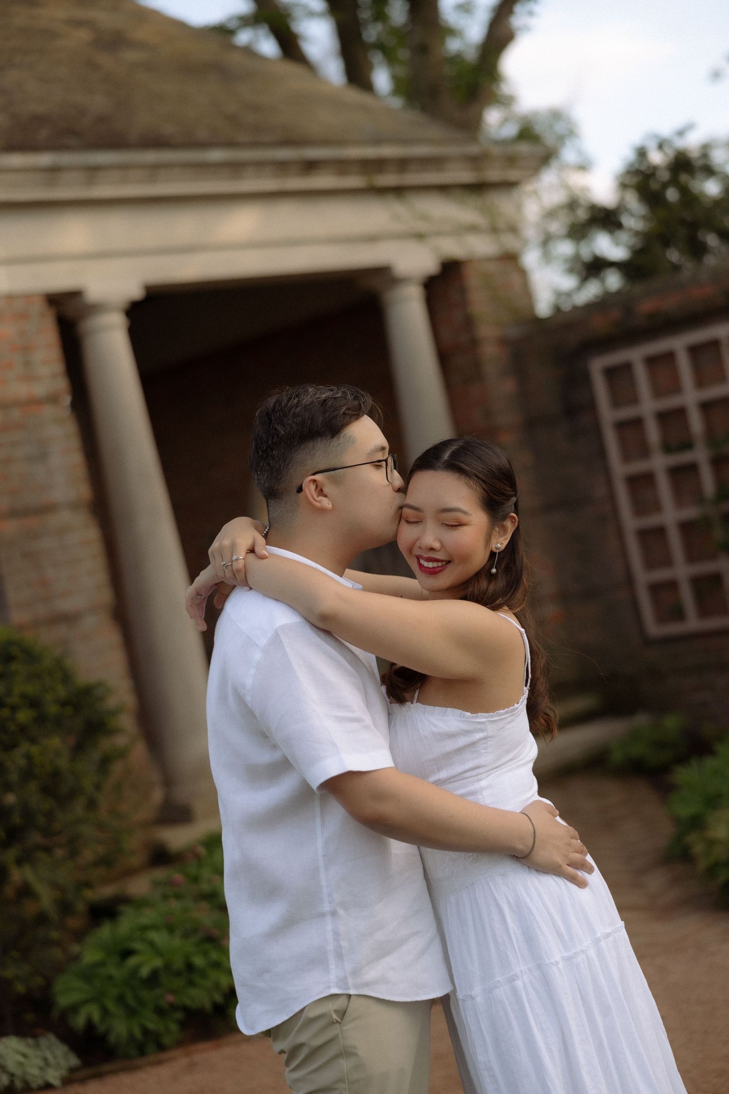 A couple in white outfits embracing and sharing a kiss outdoors near a brick building with columns.