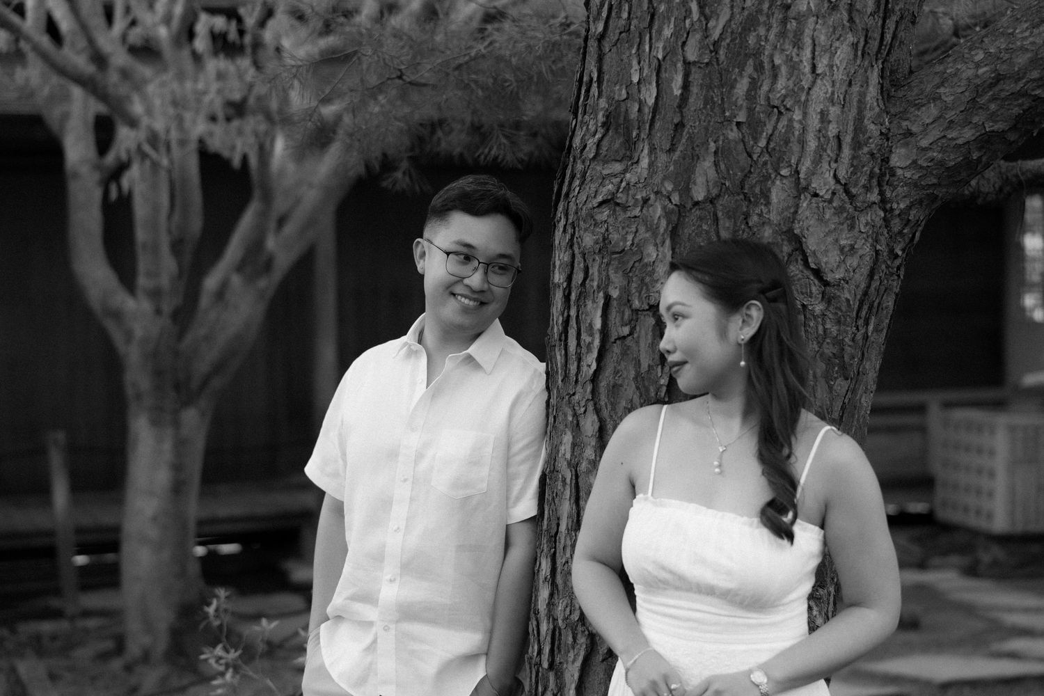 Black and white photo of a smiling couple leaning against a large tree outdoors.
