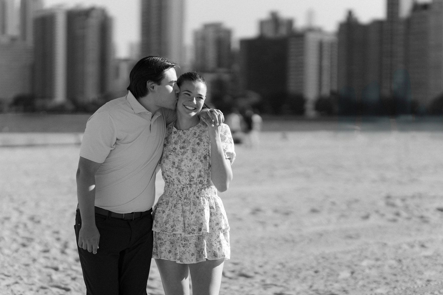 Black and white photo of couple walking on Chicago beach, man kissing woman on cheek with skyline behind.