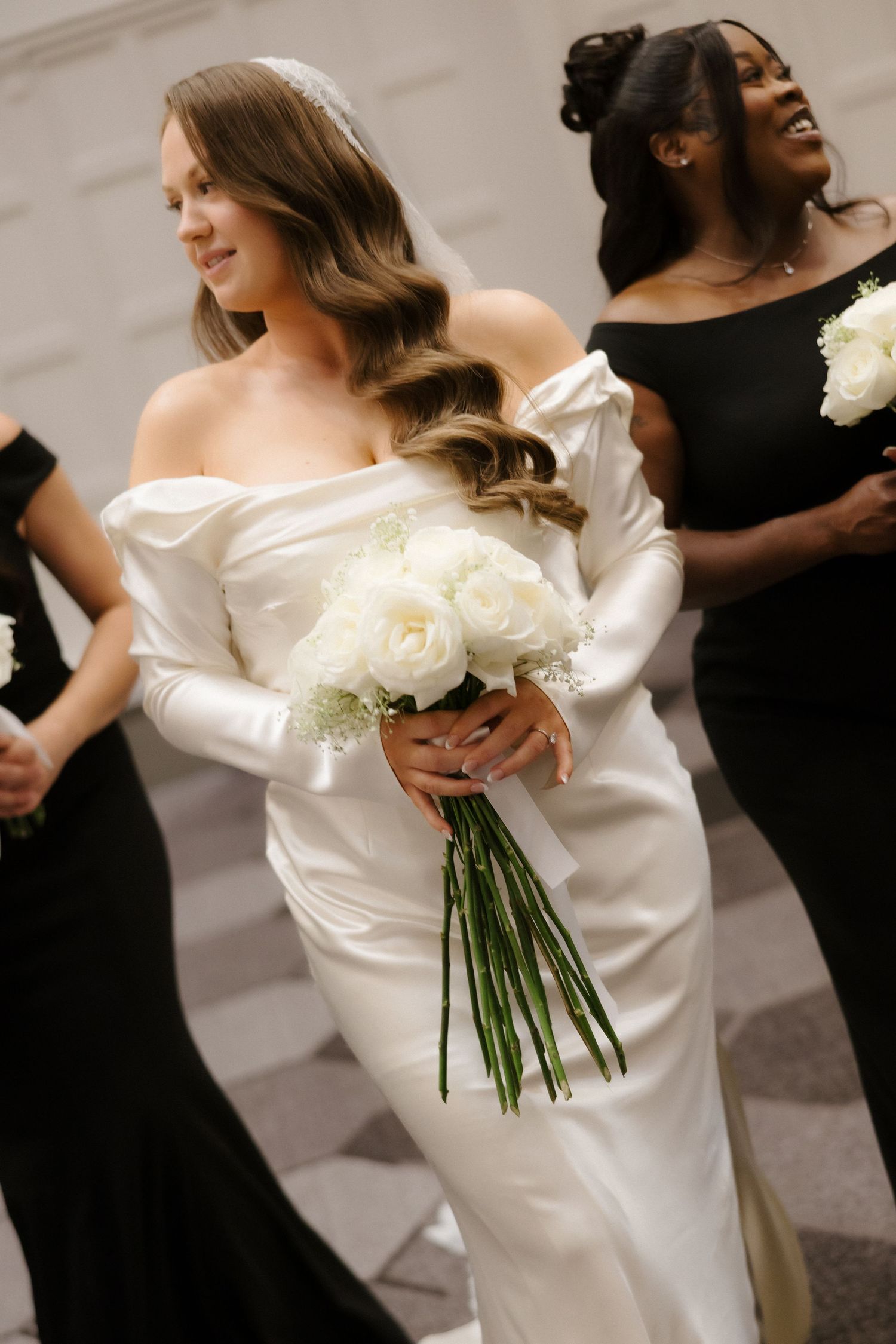 Bride in off-shoulder white satin gown holds white rose bouquet, flanked by bridesmaids in black dresses.