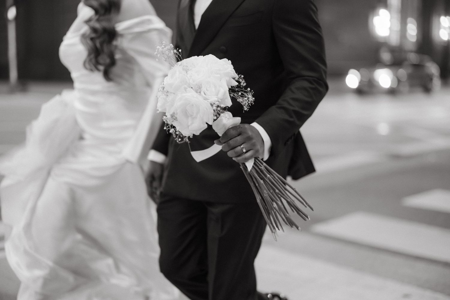 Black and white photo of bride and groom walking, groom holding white rose bouquet outdoors.