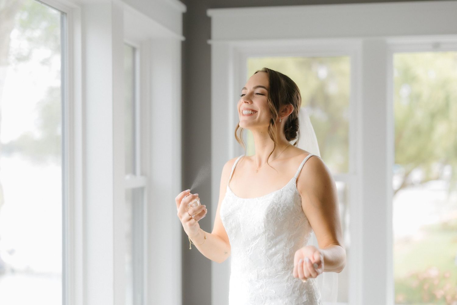 Smiling bride in white lace wedding gown standing near bright windows on her wedding day.