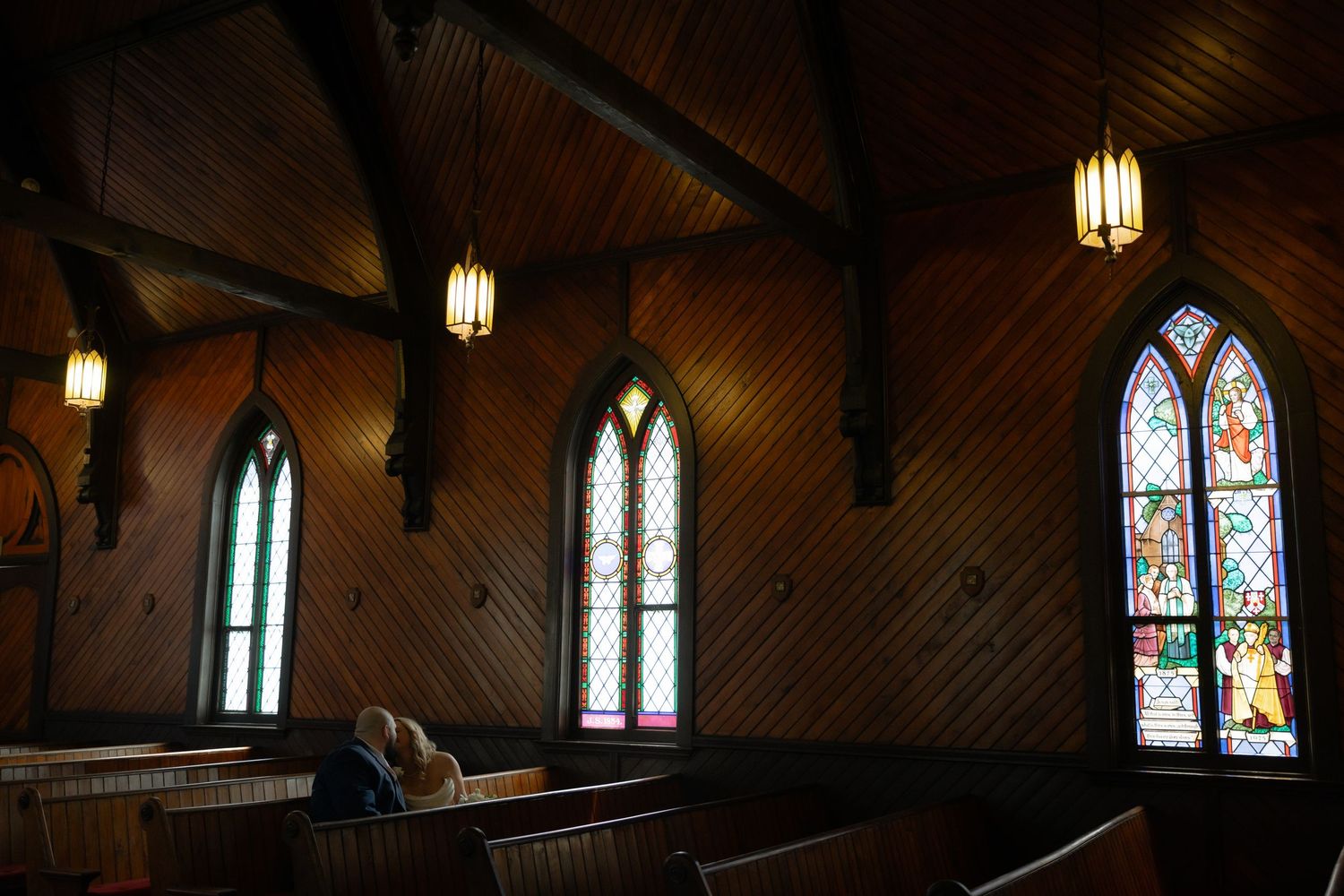 Historic wooden church interior with colorful stained glass windows, hanging lanterns, and oak pews in warm lighting.