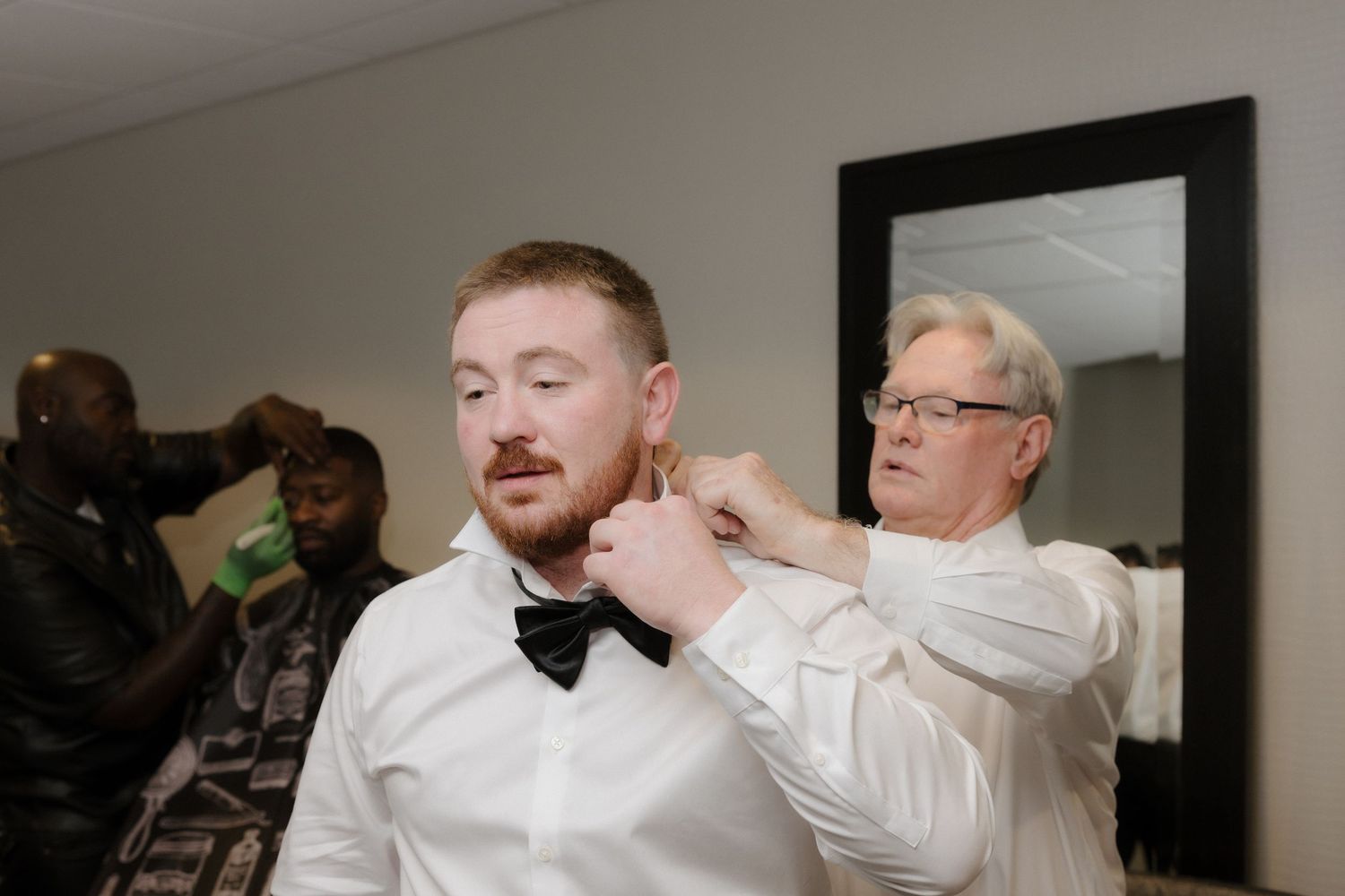 A groom in a white shirt gets his black bow tie adjusted by an older man in glasses before a wedding ceremony.