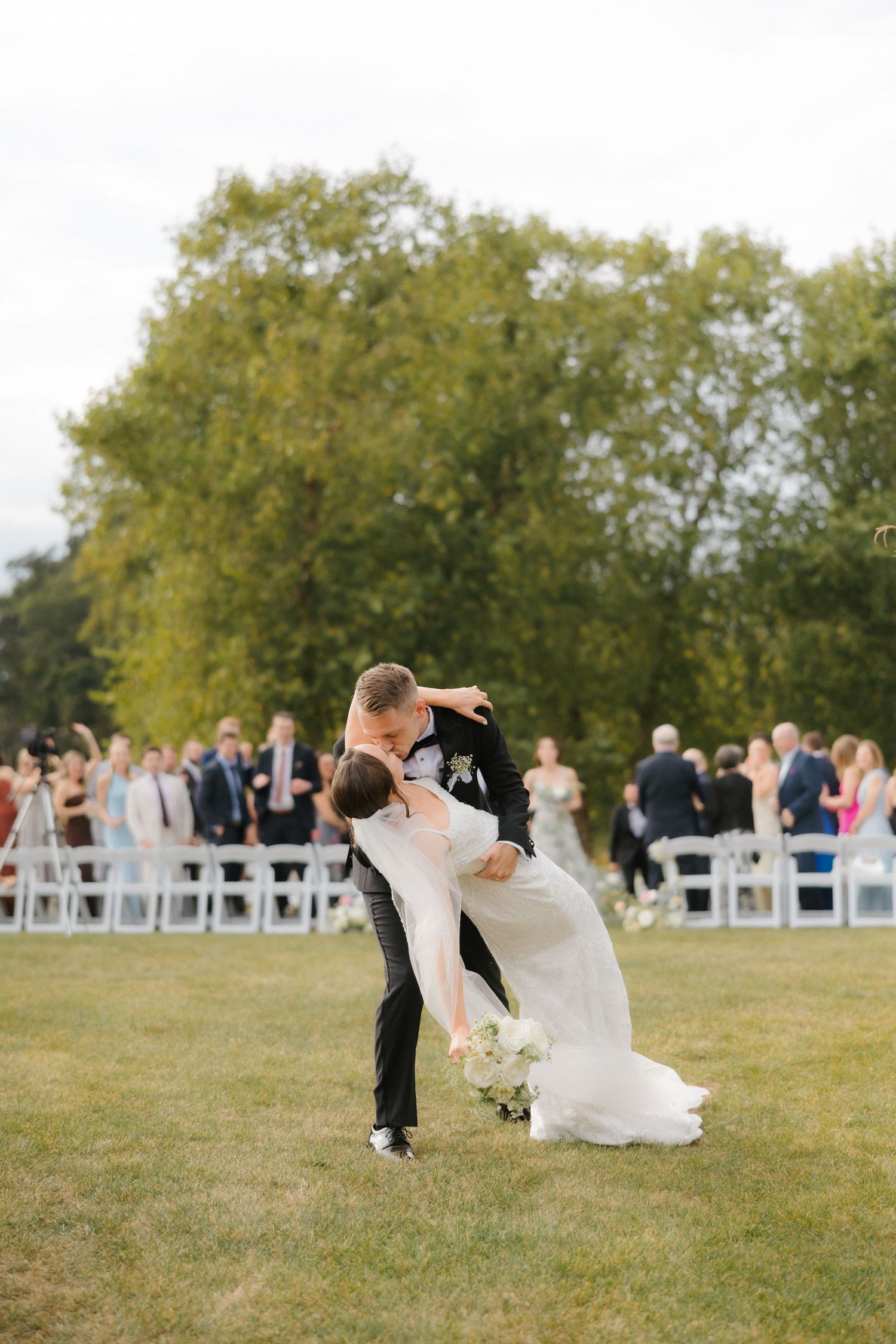 Groom dips bride for a romantic kiss on green lawn after outdoor wedding ceremony with guests seated in white chairs.