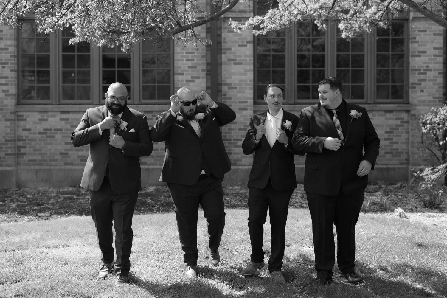 Black and white photo of four groomsmen in suits adjusting their ties and boutonnieres outdoors near a brick building.