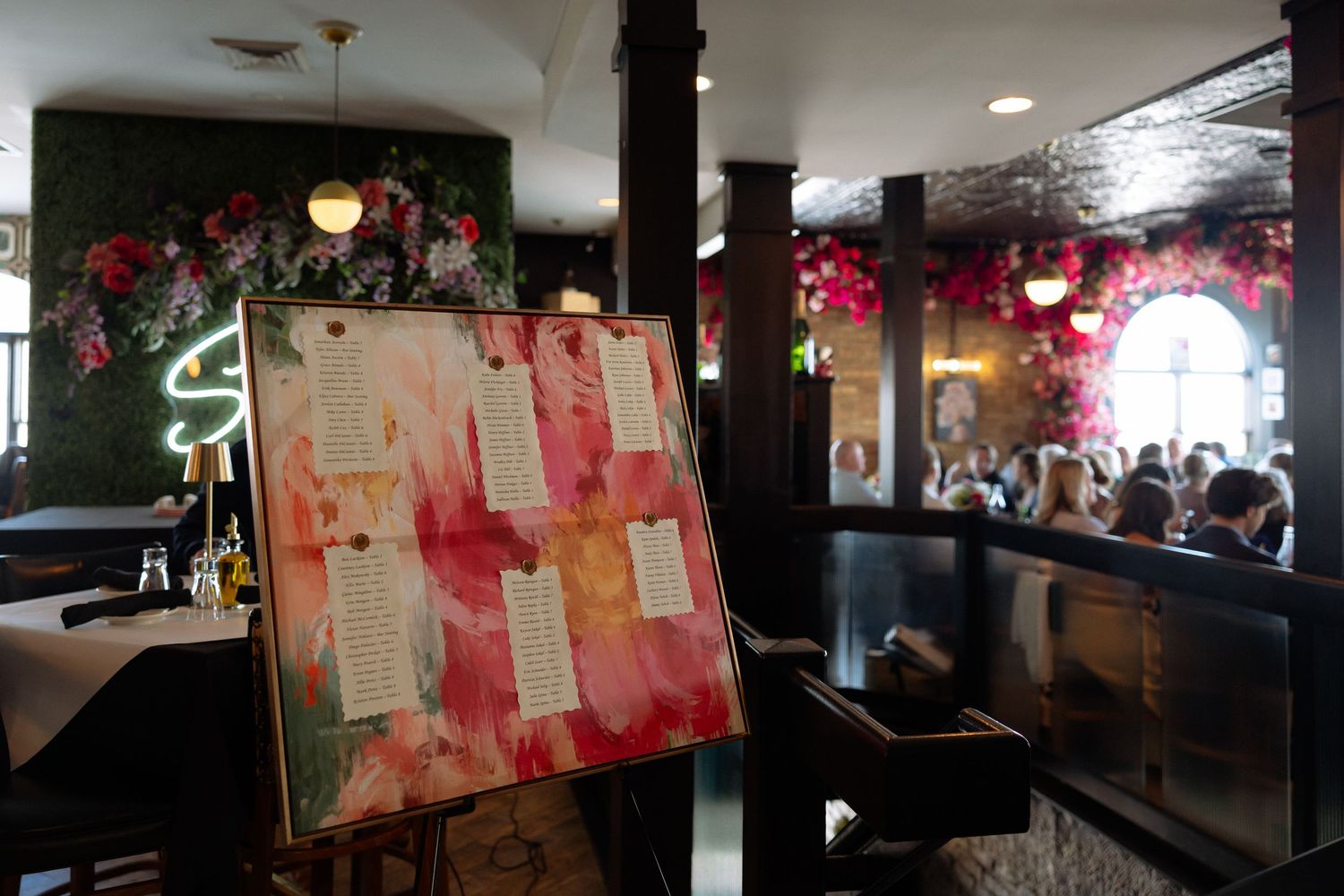 Wedding seating chart on easel with pink floral decor in a busy reception venue.