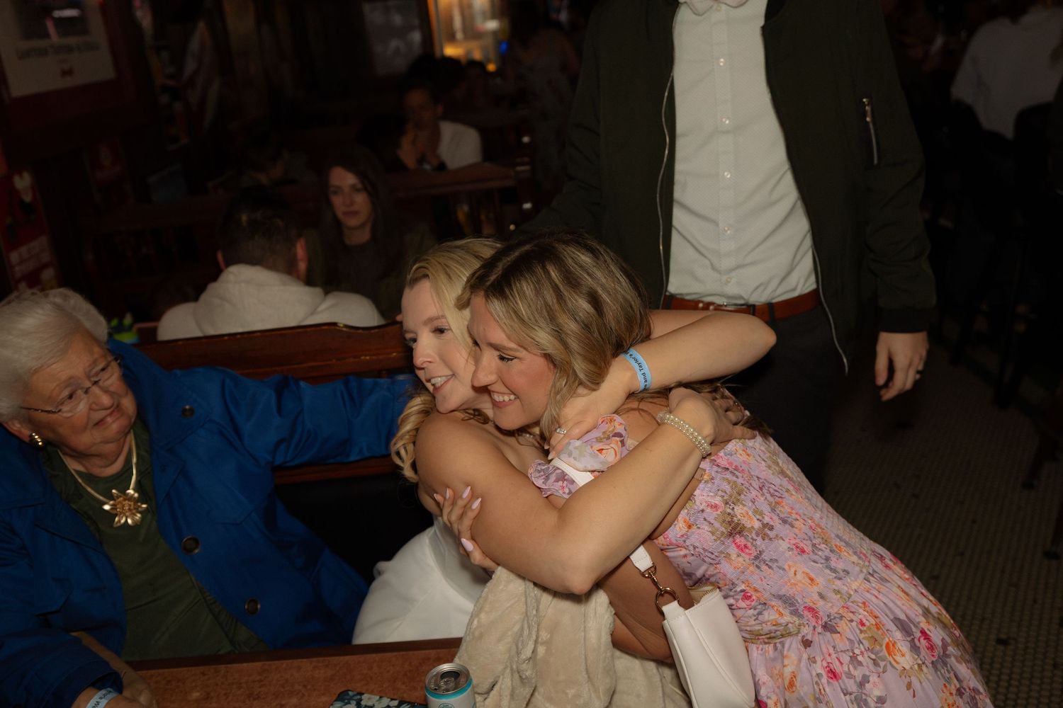 Two women sharing a warm hug in a cozy restaurant while an elderly woman in blue sits nearby.