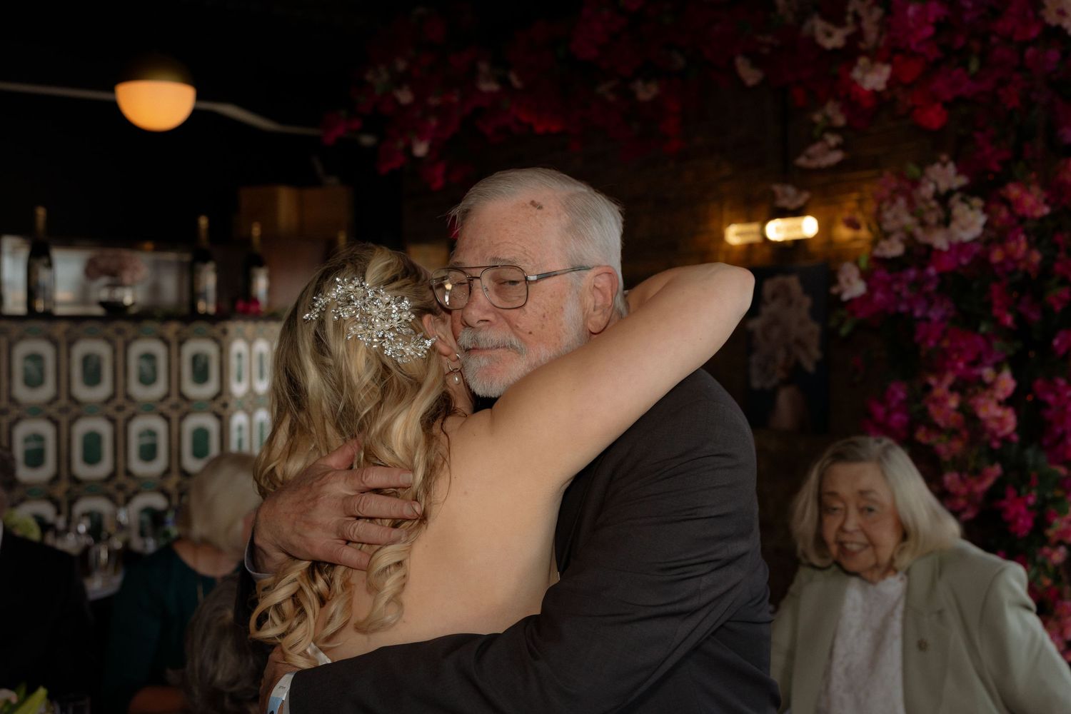 Elderly man in glasses sharing a warm hug with a blonde bride in a backless gown at a floral wedding reception.