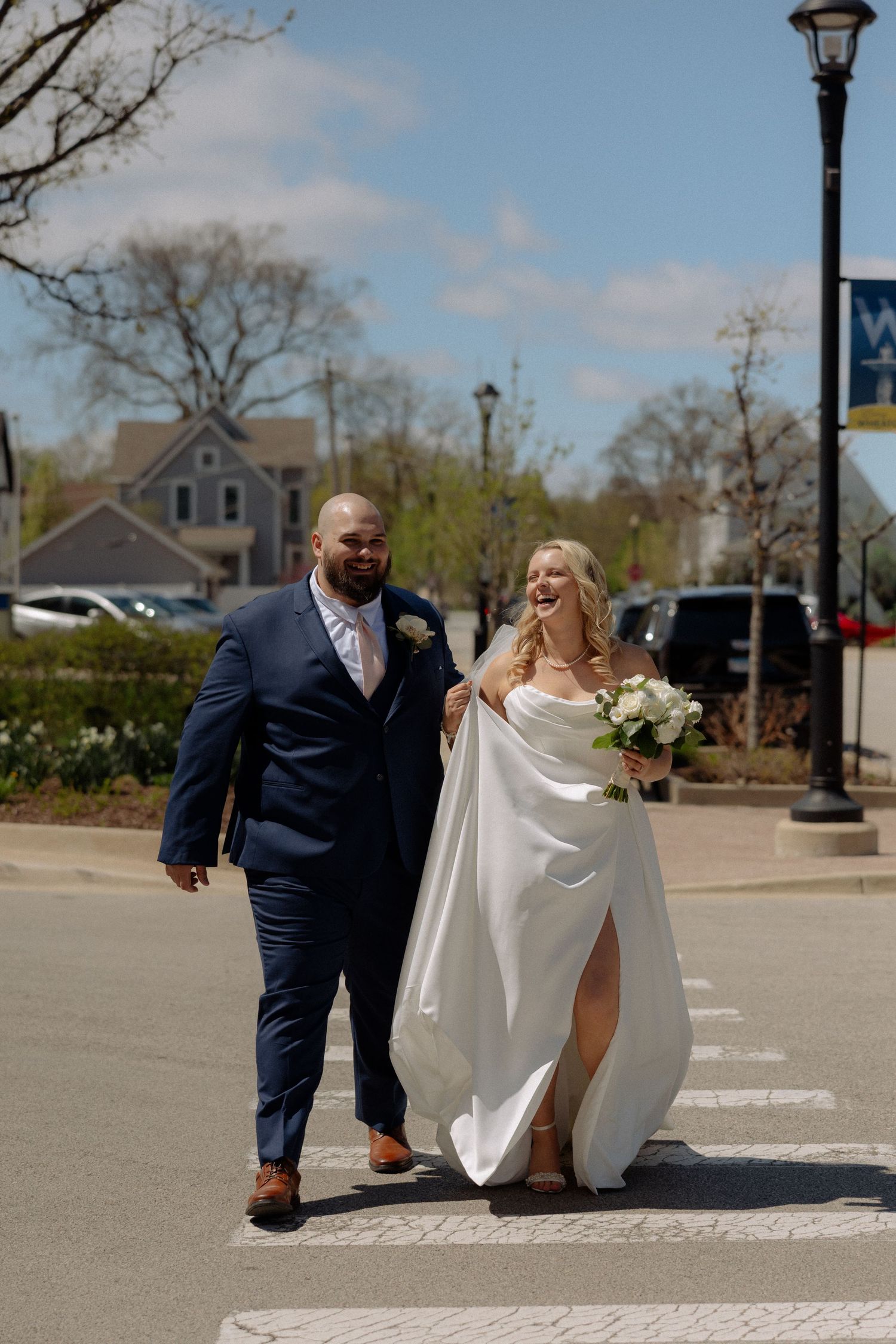 A bride and groom walk hand in hand across a crosswalk on a sunny day, she in a white gown, he in a navy suit.