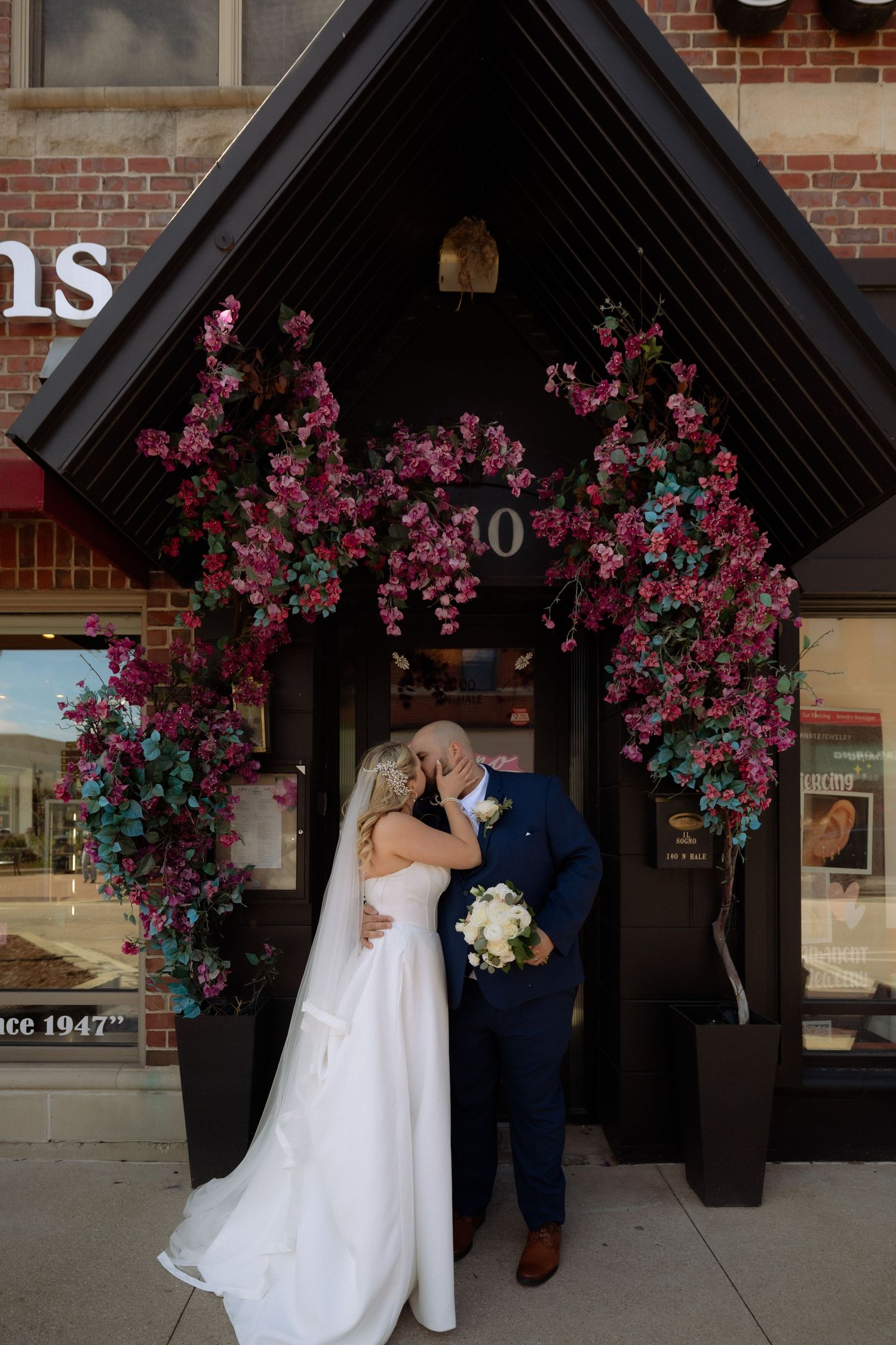 Bride and groom kissing under a floral arch with pink blooms at a brick building entrance.