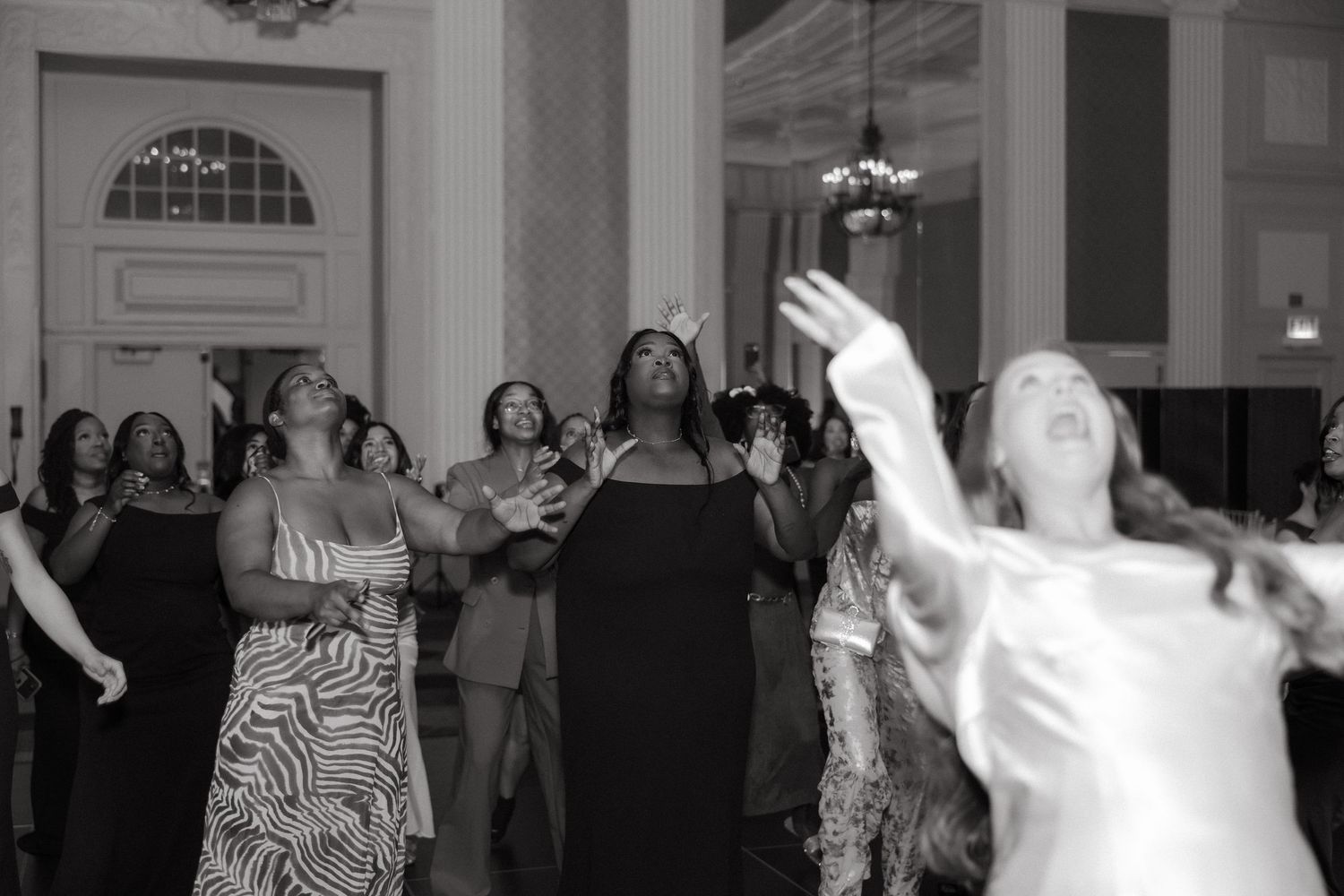 Black and white photo of women reaching up at a wedding bouquet toss in an elegant ballroom.