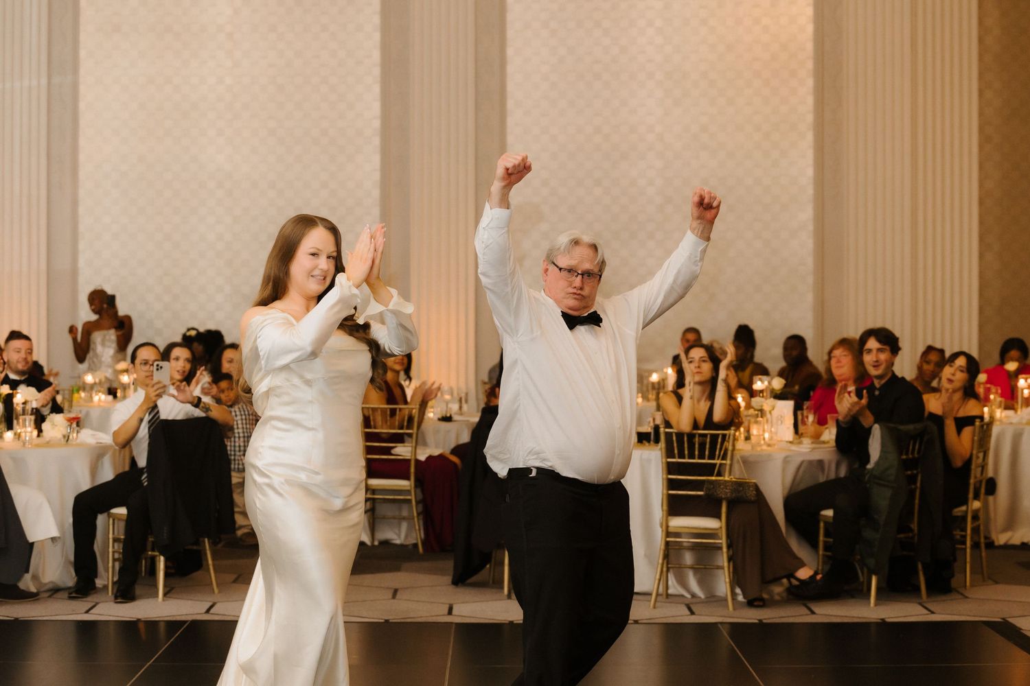 Bride and father joyfully raise their arms during a wedding reception first dance in an elegant ballroom.