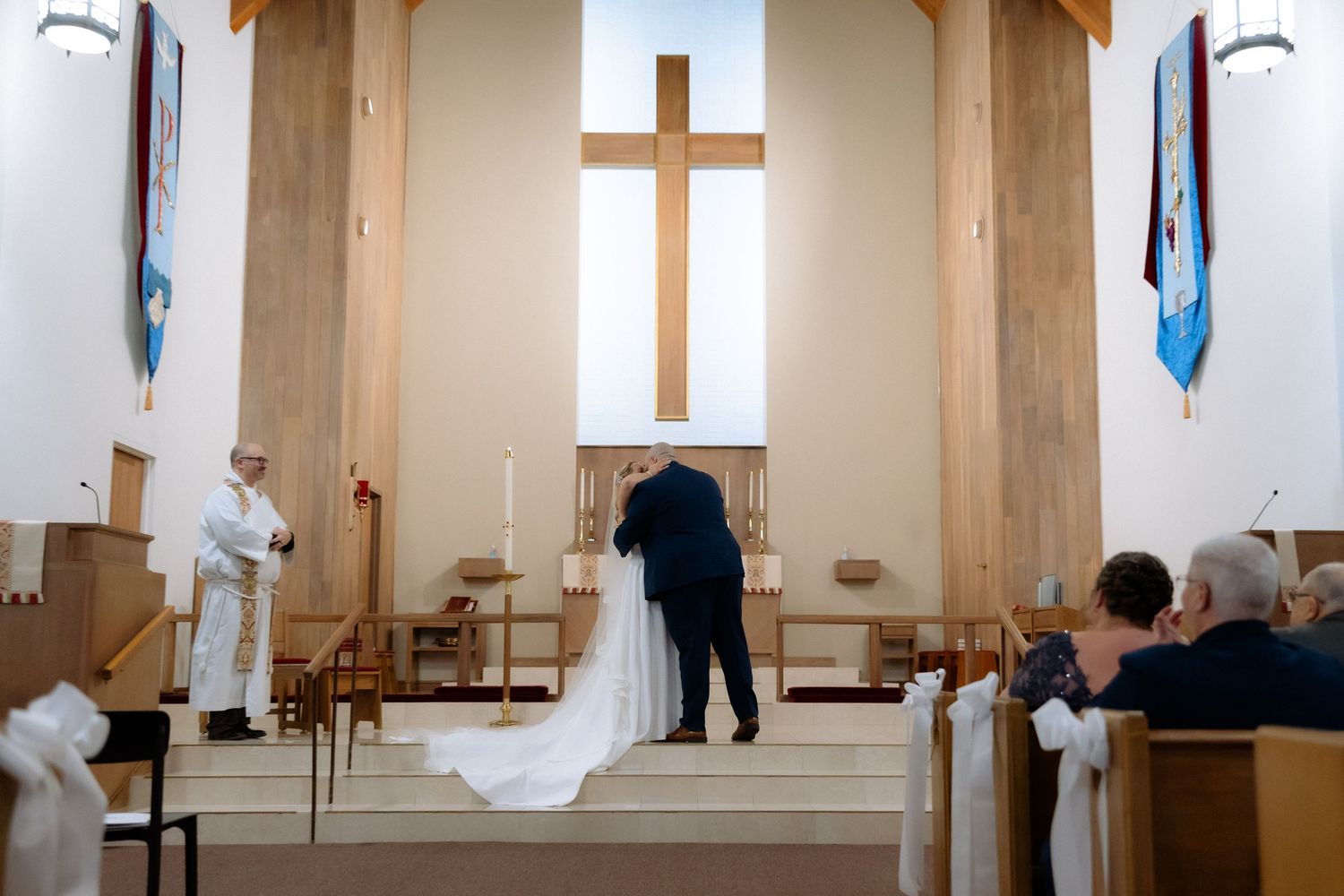 A bride and groom exchange vows at a church altar with a large wooden cross, as an officiant and guests witness the ceremony.