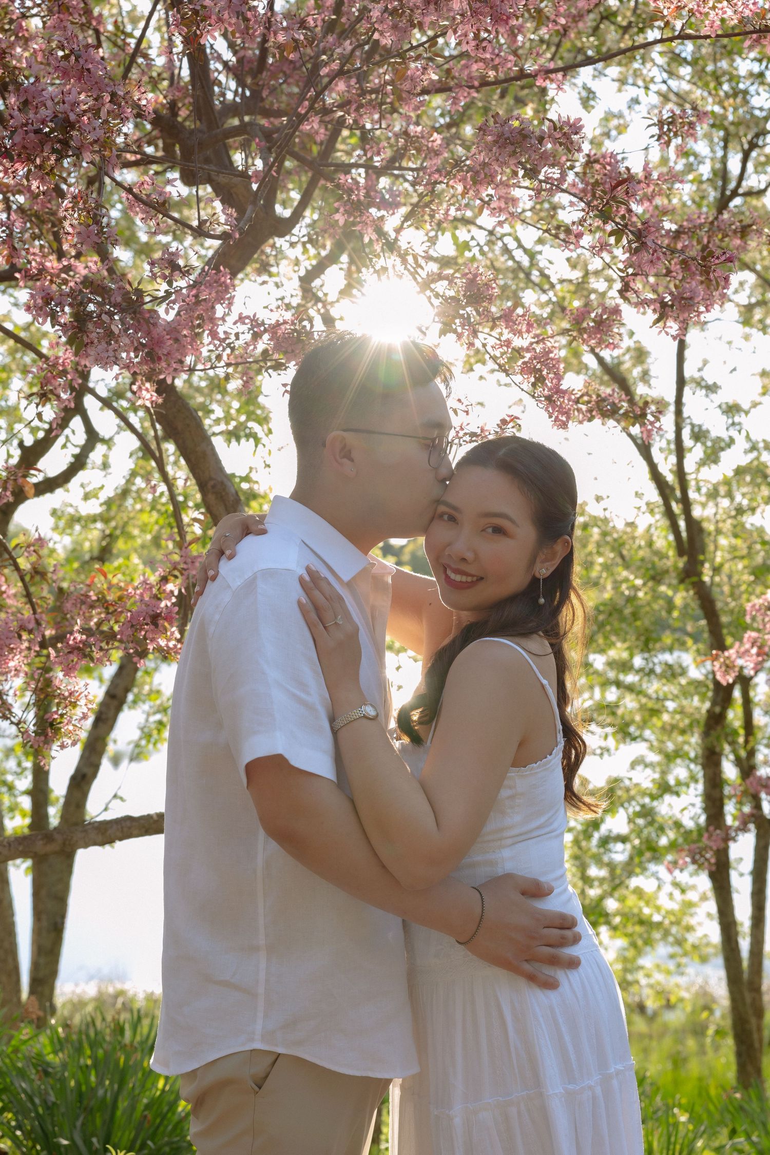 Couple embracing under pink cherry blossom trees in golden sunlight, woman in white dress smiling at camera.